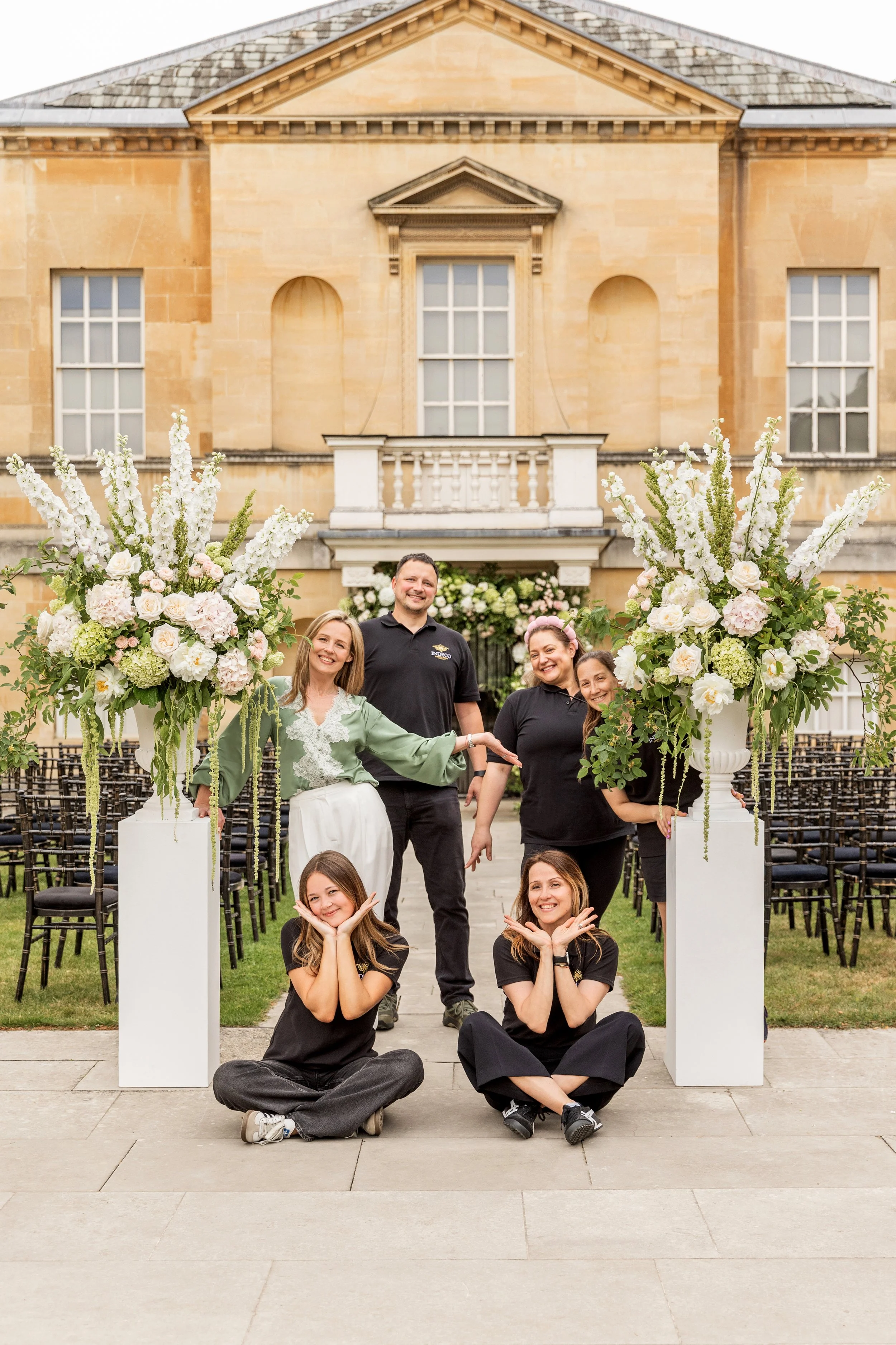 A team of florists posing in an outdoor wedding or event setting with floral arrangements and a historic Chiswick building in the background.