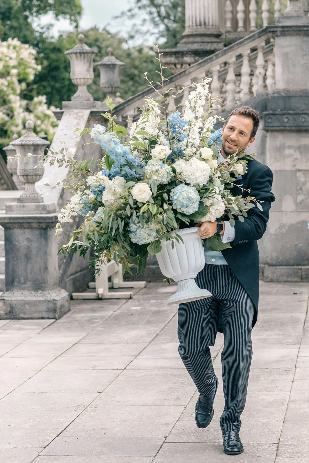 Member of the wedding party carrying a large white and blue floral urn arrangement at Chiswick House & Gardens.