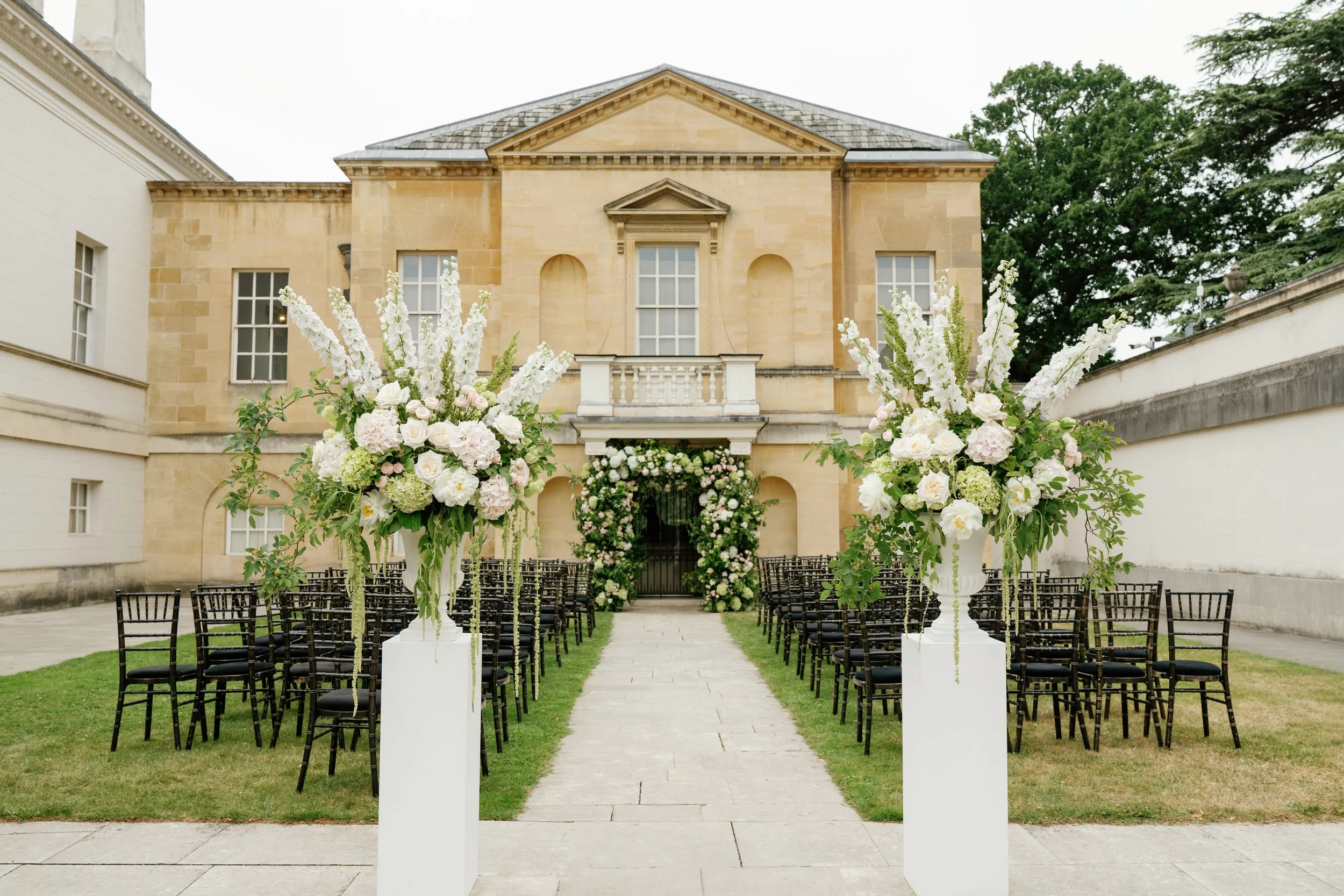 Outdoor wedding ceremony setup with black chairs arranged in rows on a grassy lawn, leading to a historic building with a beige stone facade. There are large floral arrangements on white pedestals lining the aisle, featuring white and blush pink flowers. The setting is surrounded by tall trees.