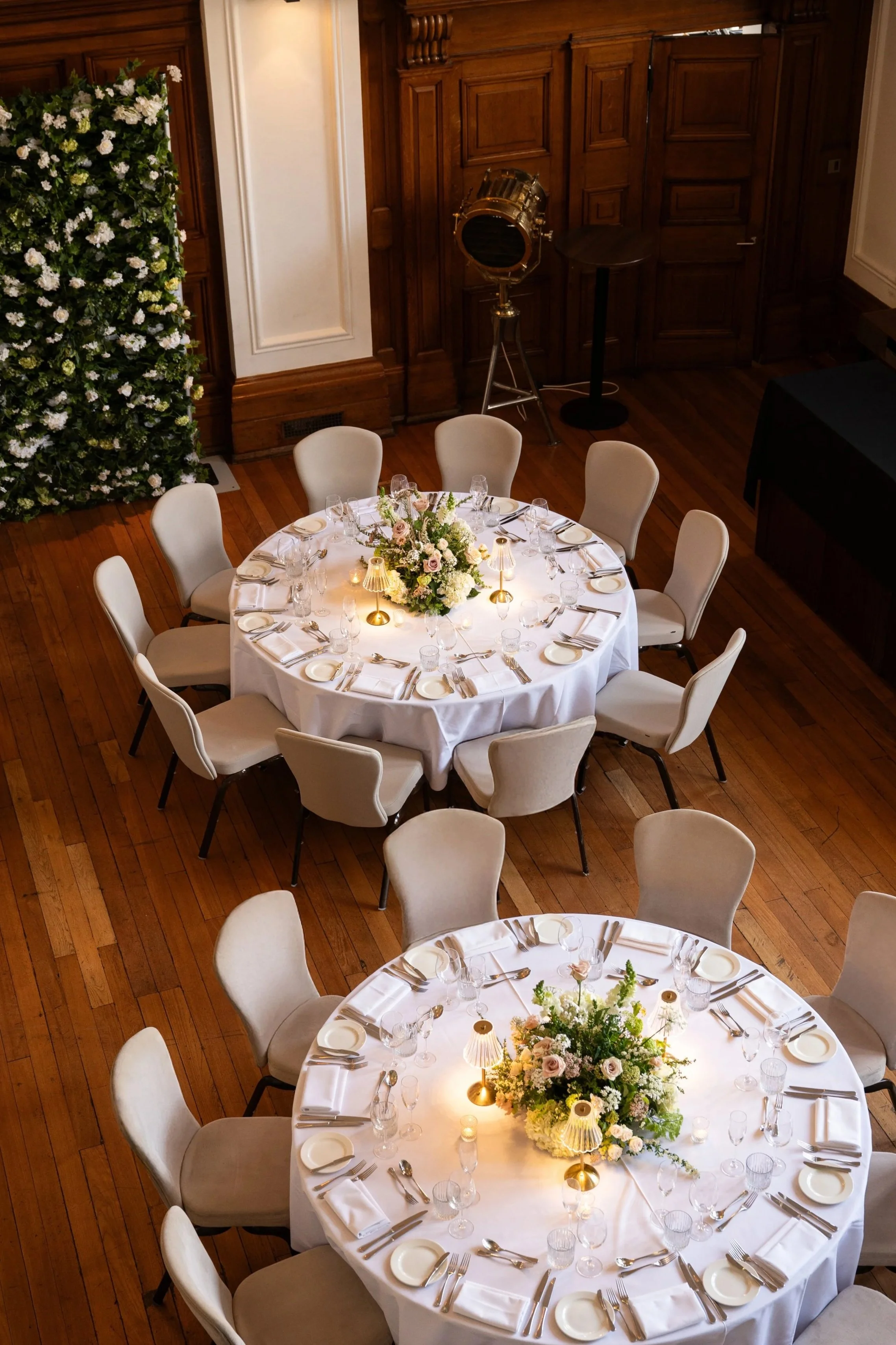A round banquet table set for a wedding or formal event, with a white tablecloth, floral centerpiece, and neatly arranged place settings, in a wood-paneled room.