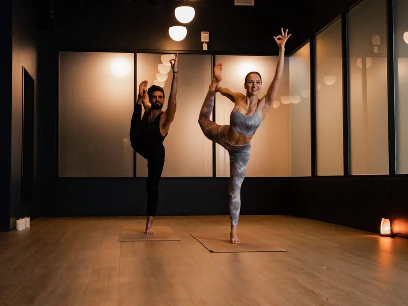 Deux personnes pratiquent le yoga dans un studio, en posant une posture d'équilibre sur une jambe, avec une ambiance chaleureuse et une lumière tamisée.
