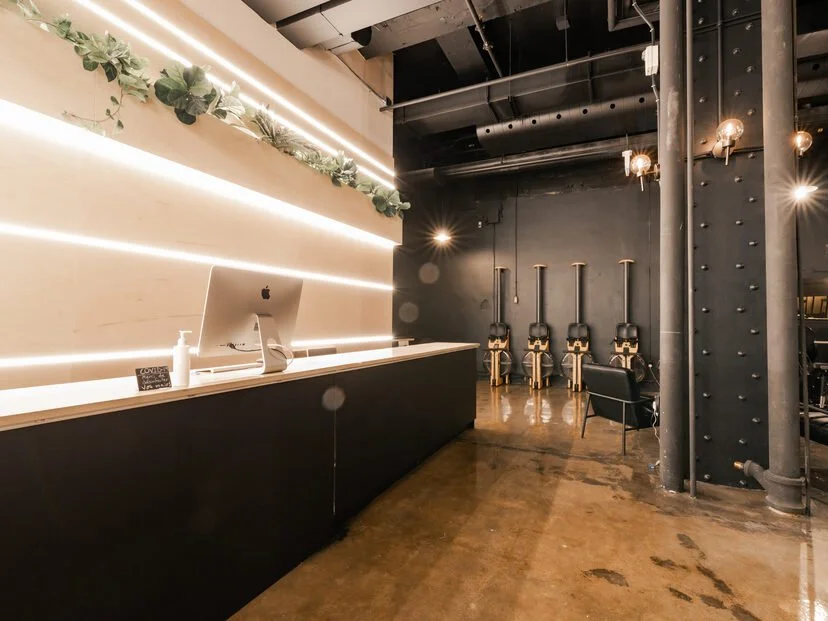 Un comptoir de réception avec un ordinateur tout en haut à gauche, décoré avec des plantes vertes, dans un intérieur moderne. En arrière-plan, des machines à coiffure ou à barbier alignées contre un mur noir, avec un fauteuil noir à proximité.