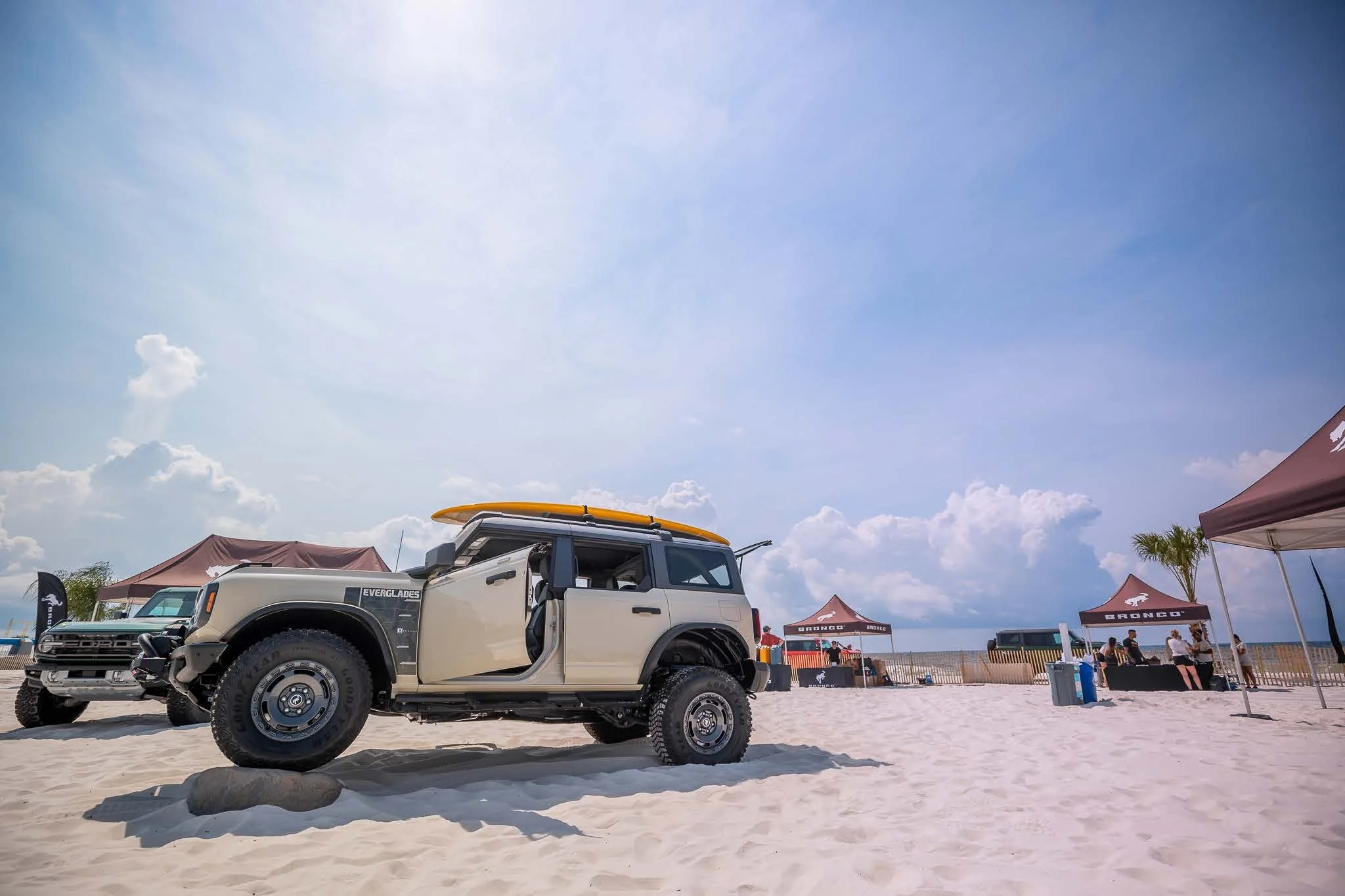 Ford Bronco SUVs displayed on a sandy beach at an outdoor event with branded tents and a bright blue sky