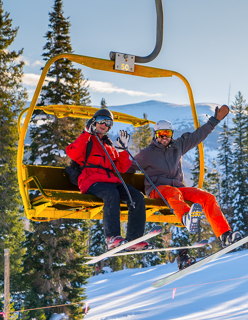 Two skiers riding a yellow chairlift above a snowy mountain slope, wearing helmets and ski goggles.