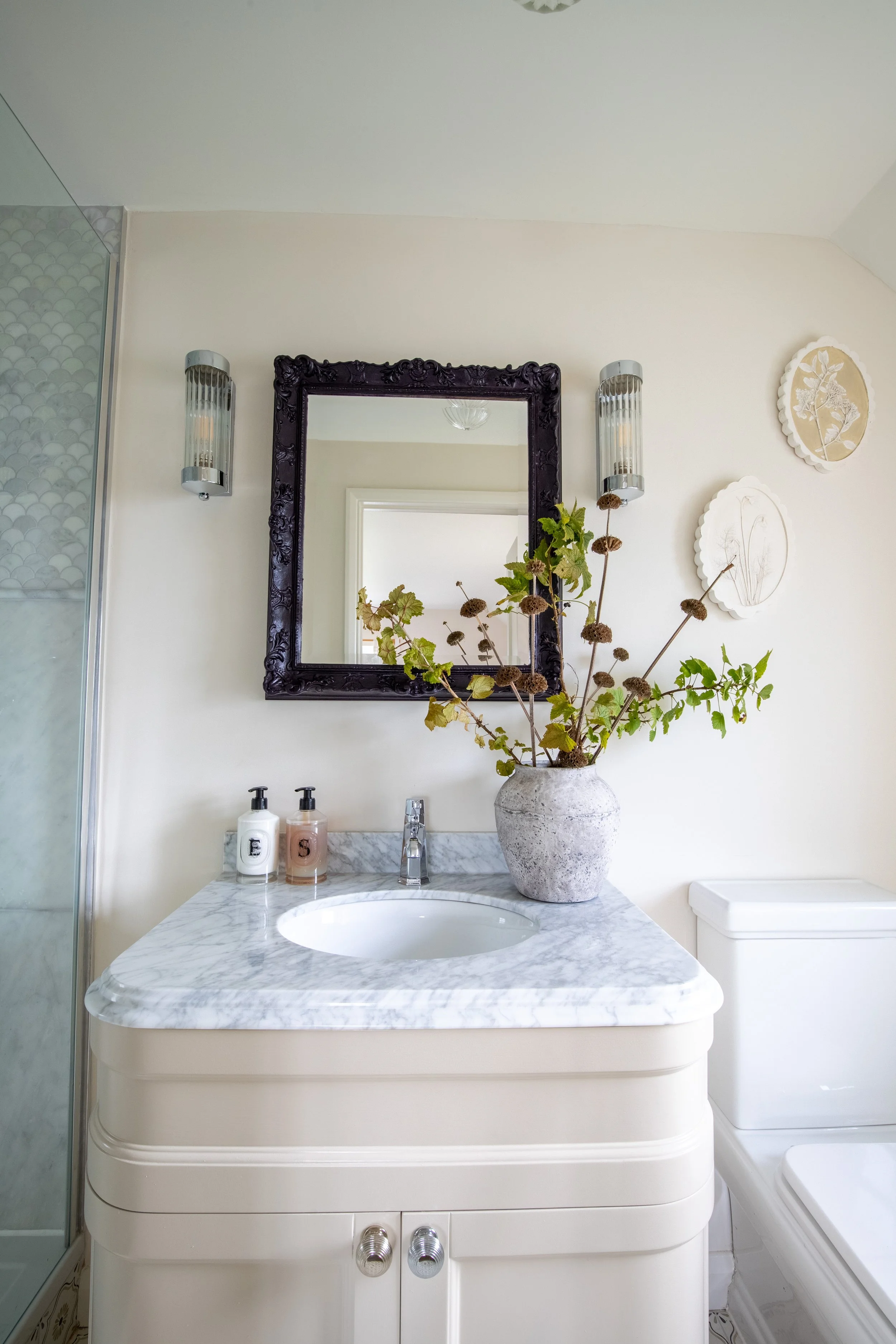 Bathroom vanity with marble countertop, a vase with dried flowers, two soap dispensers, a mirror, and wall fixtures.
