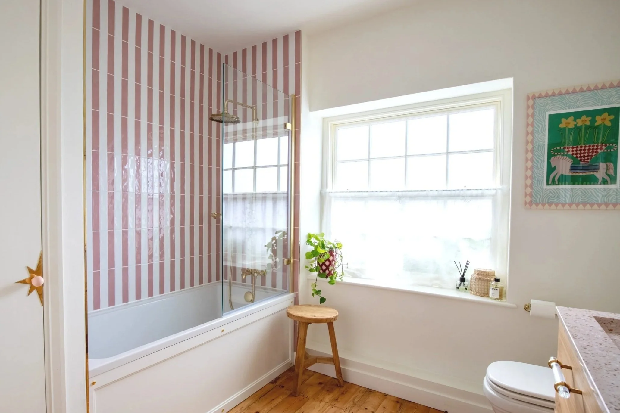 Bathroom featuring a bathtub with pink and white striped tile behind it, a glass shower door, a wooden stool next to a window, and a white toilet.