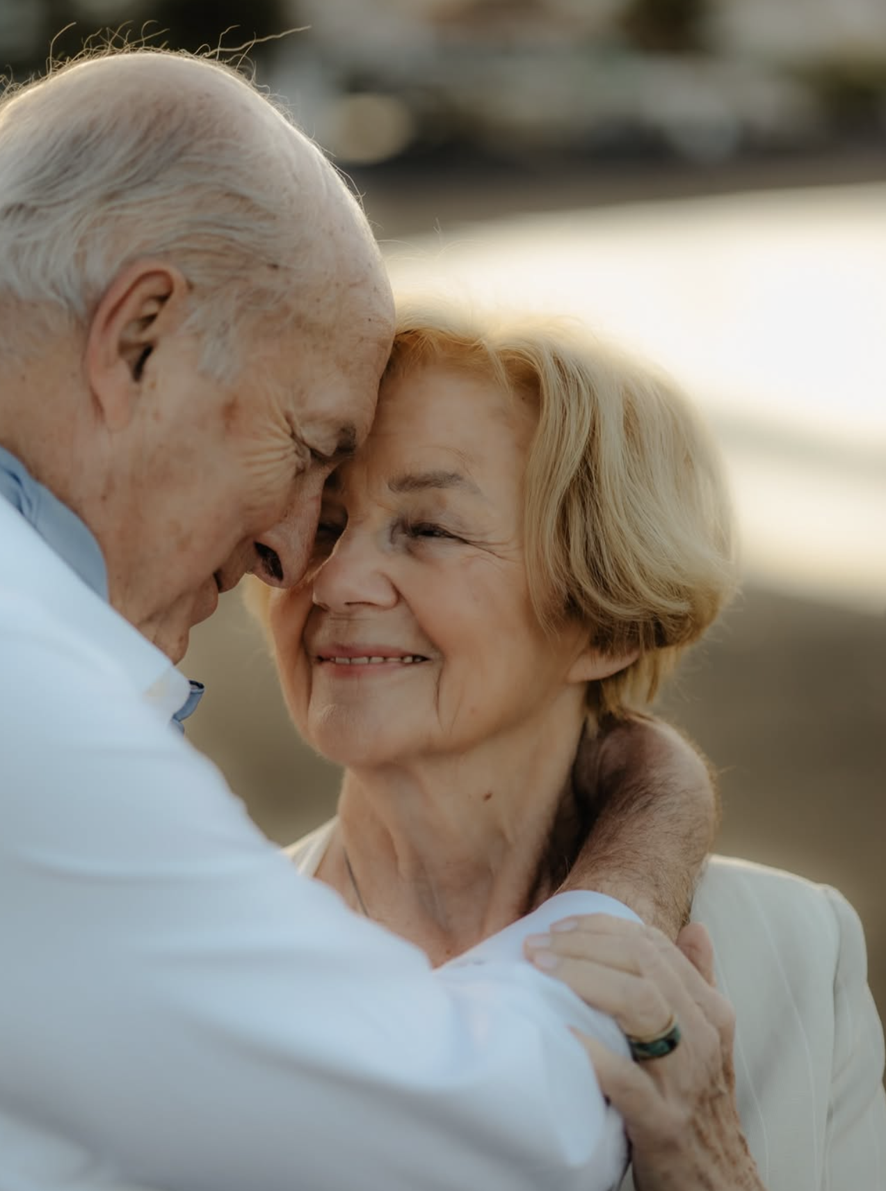 Un homme âgé et une femme âgée partagent un moment d'affection, se tenant la tête et se regardant avec tendresse, lors d'une rencontre chaleureuse en extérieur à la lumière douce du soleil.