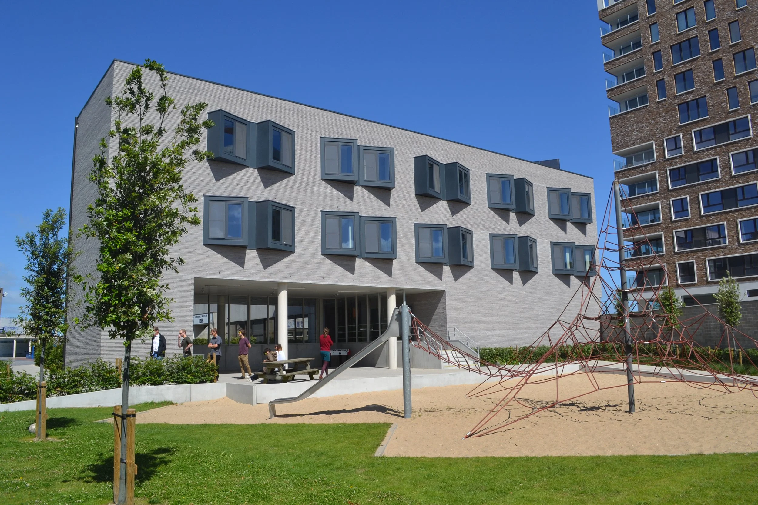 Moderne witte school of kantoorgebouw met grote, hoekige ramen, omgeven door een speelplaats met kinderen en jonge bomen, onder een heldere blauwe hemel.