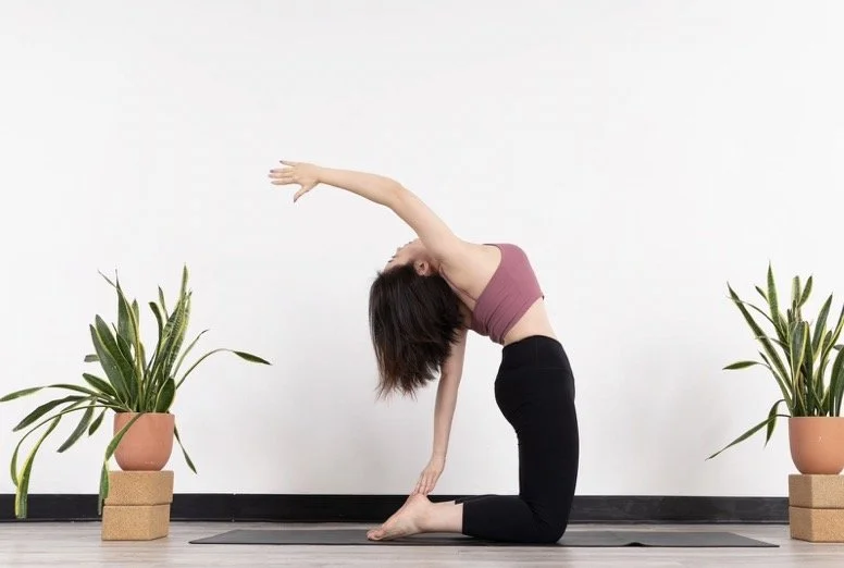 Woman practicing yoga in a kneeling forward bend stretch in a minimalist room with two potted plants.