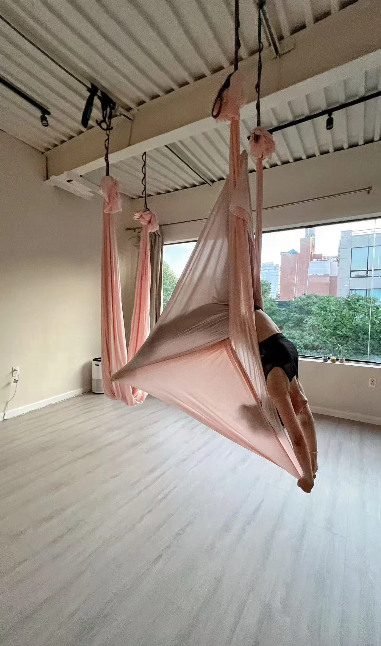 A person practicing aerial yoga in a pink hammock inside a room with large windows and a view of trees and buildings outside.