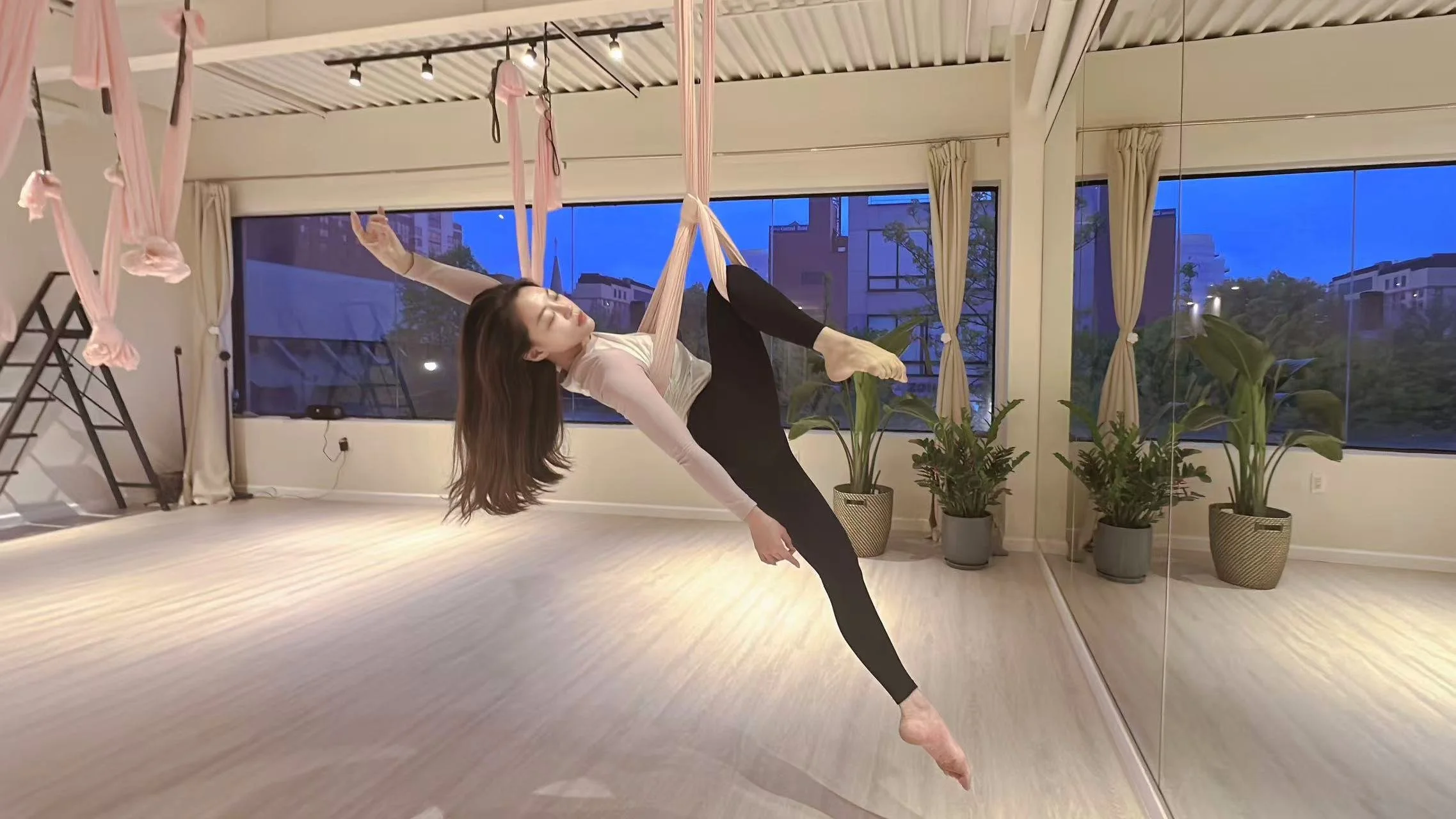 A woman practicing aerial silk yoga in a well-lit studio with large windows, potted plants, and a mirror.