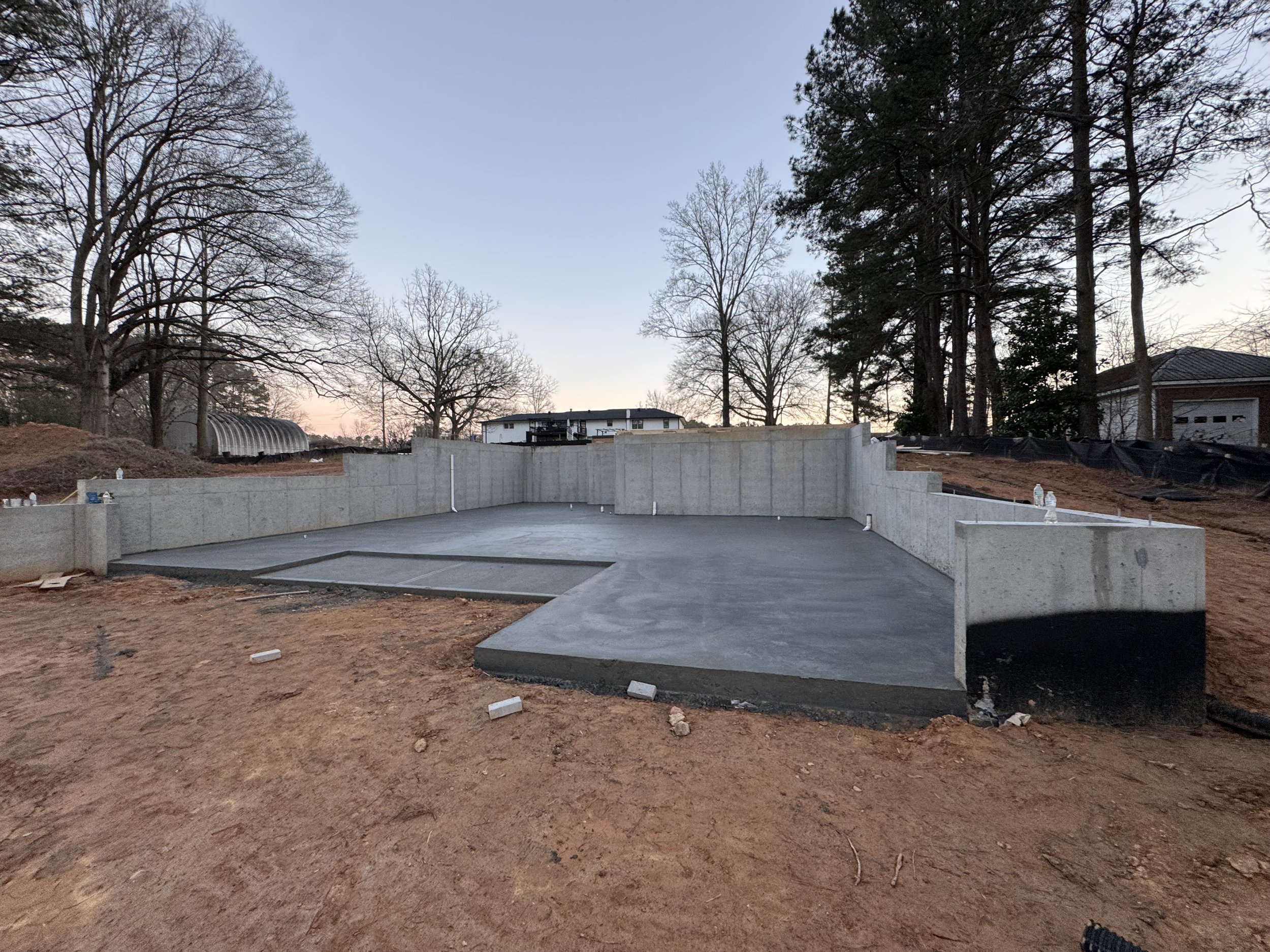 Construction site with a concrete foundation and walls, surrounded by dirt, with trees and houses in the background at dusk.