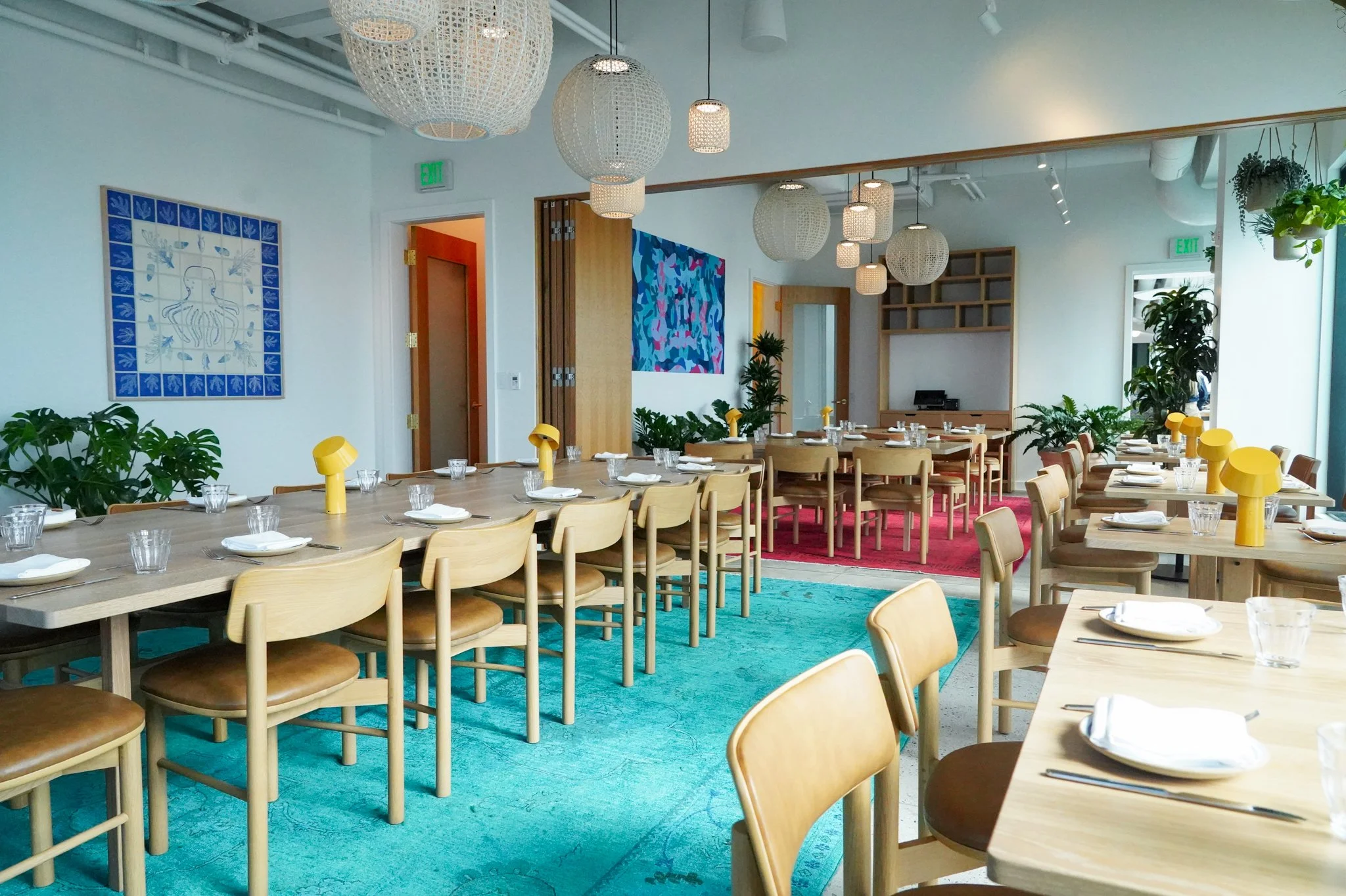 A brightly lit, modern restaurant with light wood tables and chairs, colorful artwork on the walls, woven lighting fixtures, and large windows with green plants by the window.