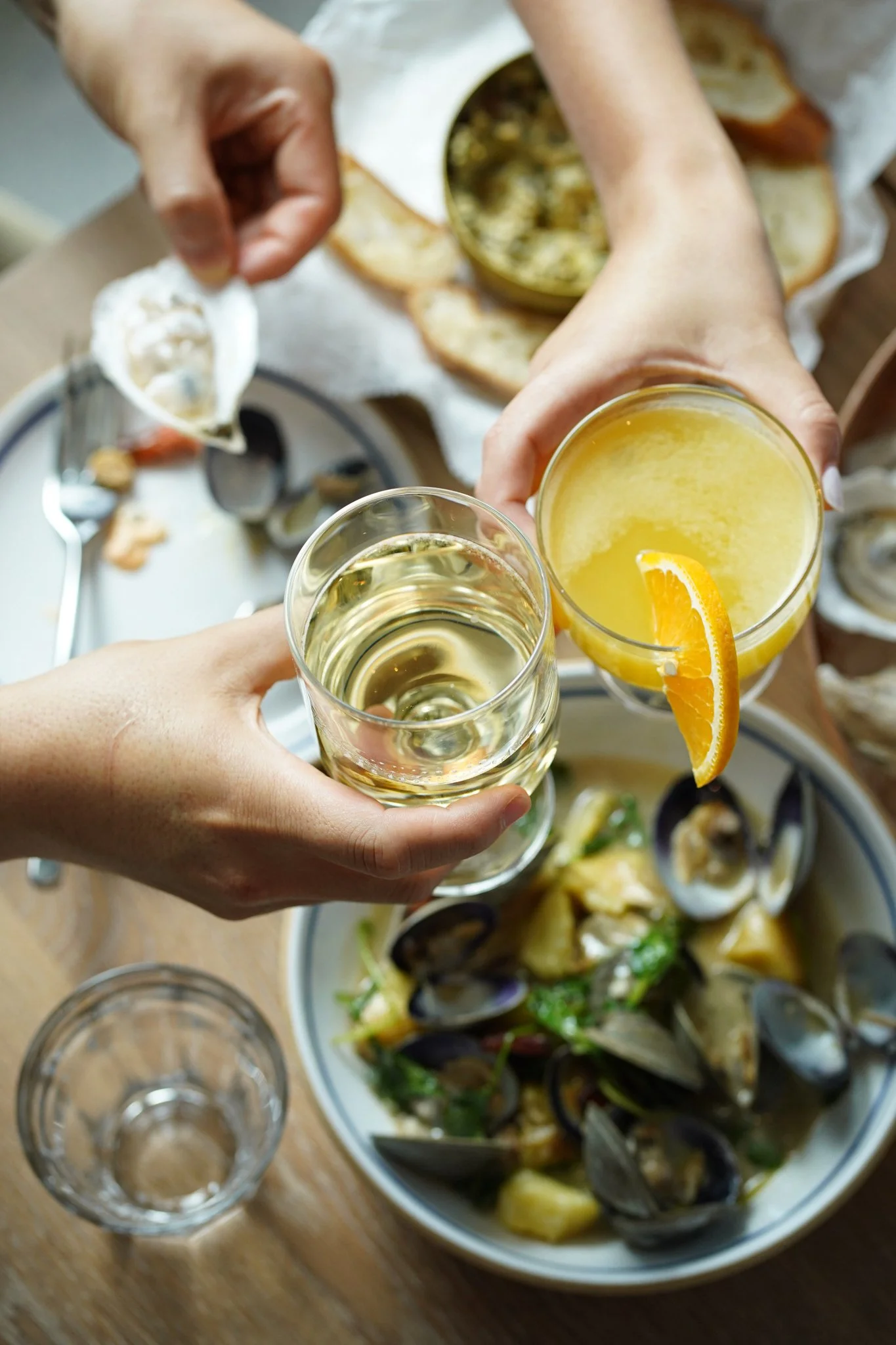 Two people clinking glasses, one with white wine and the other with a yellow cocktail garnished with an orange slice, above a table with seafood, bread, and a bowl of mussels.