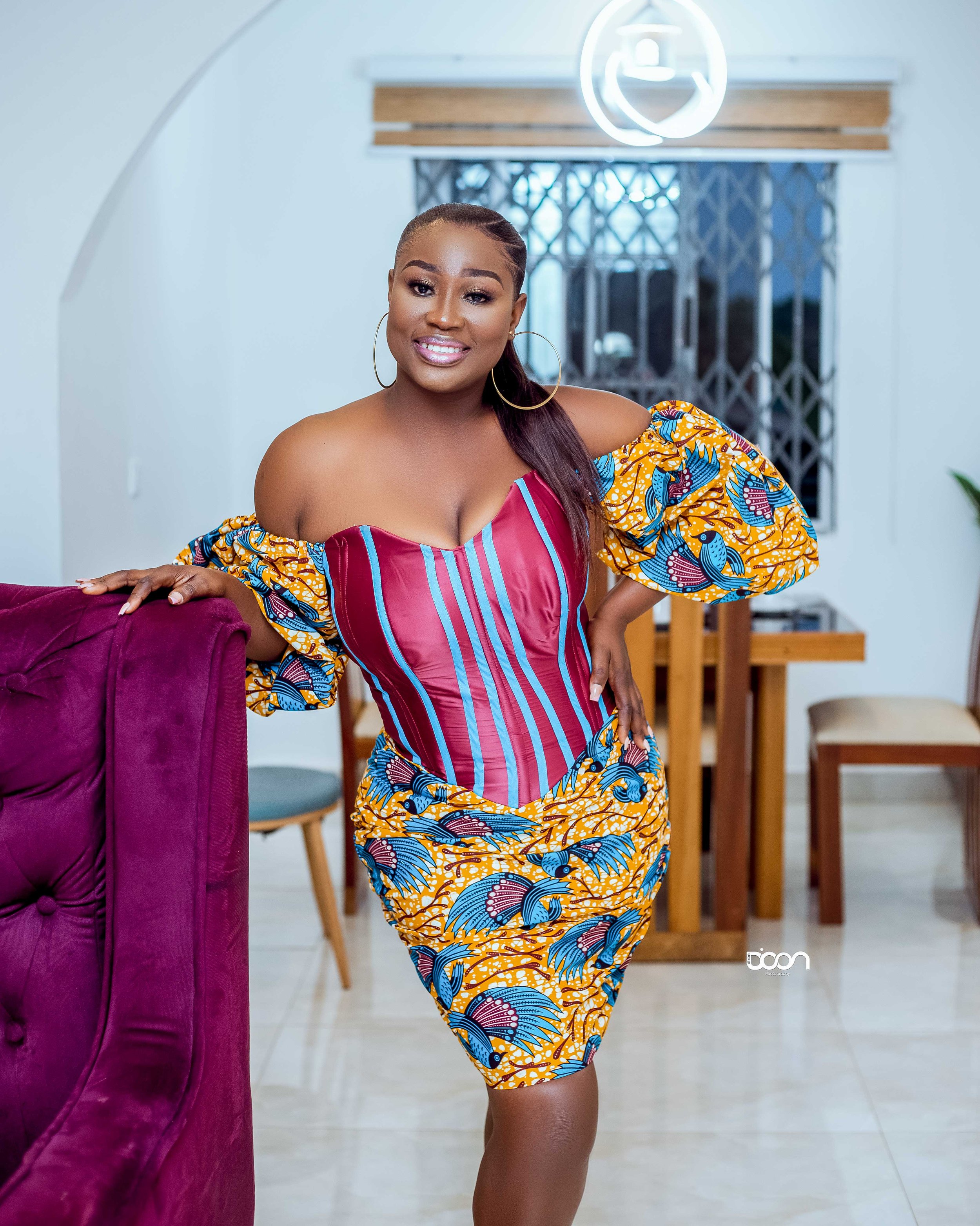 A woman in colorful traditional African attire with a patterned dress and off-the-shoulder puff sleeves, smiling and posing indoors.