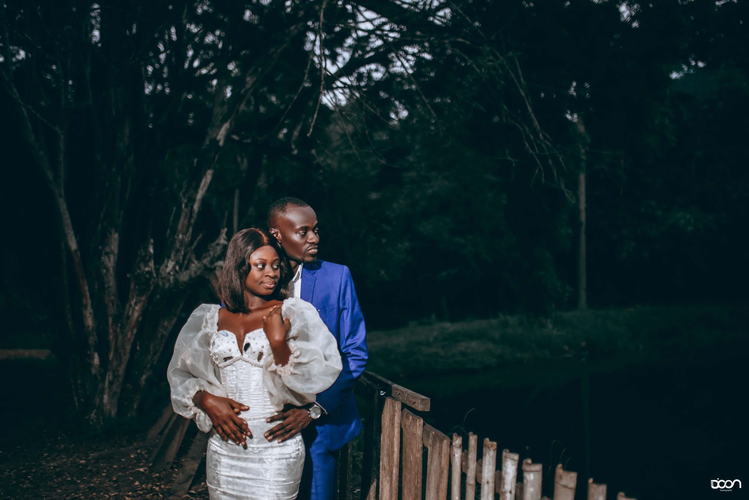 A couple dressed formally standing by a wooden railing near a body of water at night, with trees in the background.