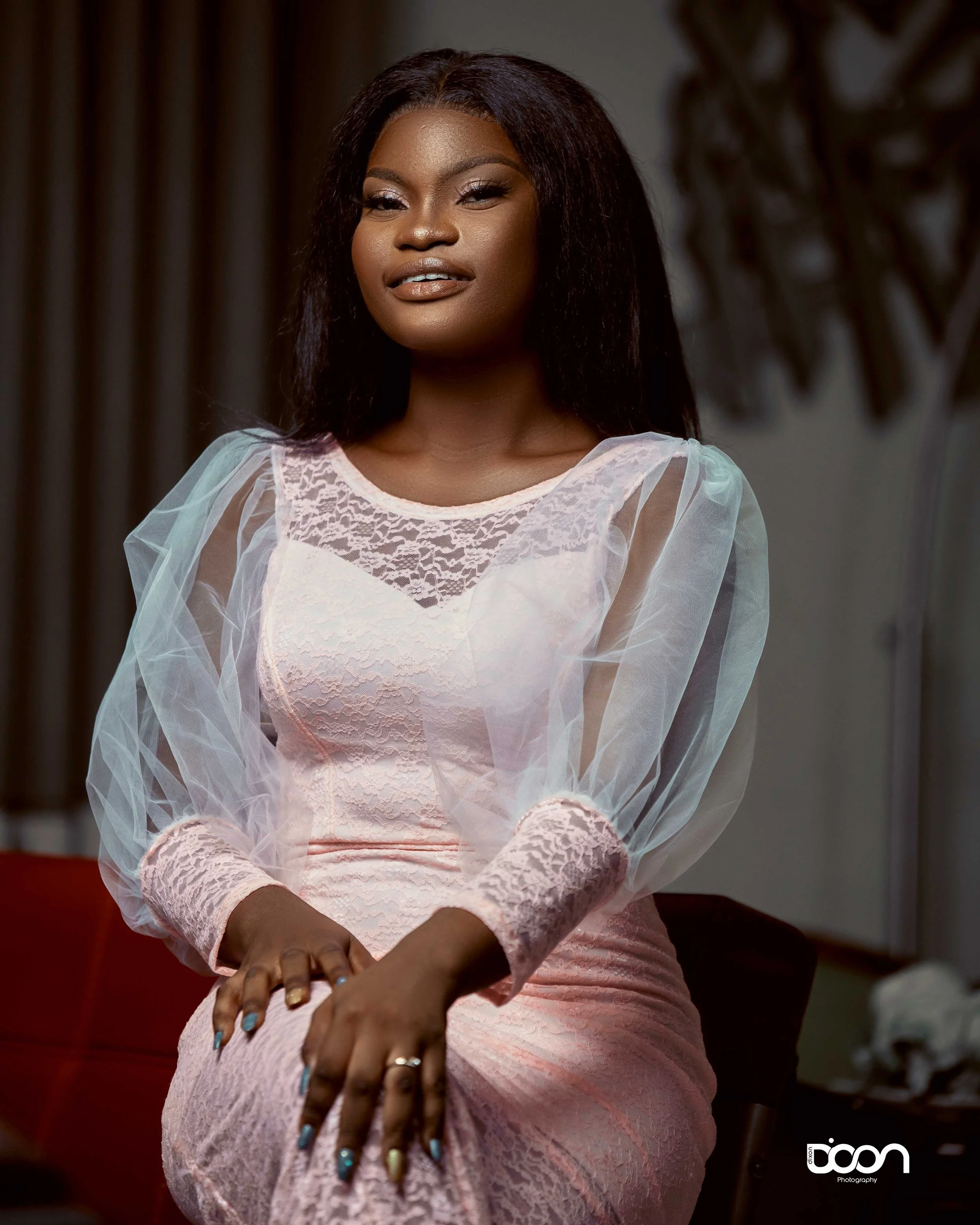A woman with long dark hair wearing a light pink lace dress and sheer puffy sleeves, sitting with her hands resting on her lap, looking slightly to the side with a soft smile.