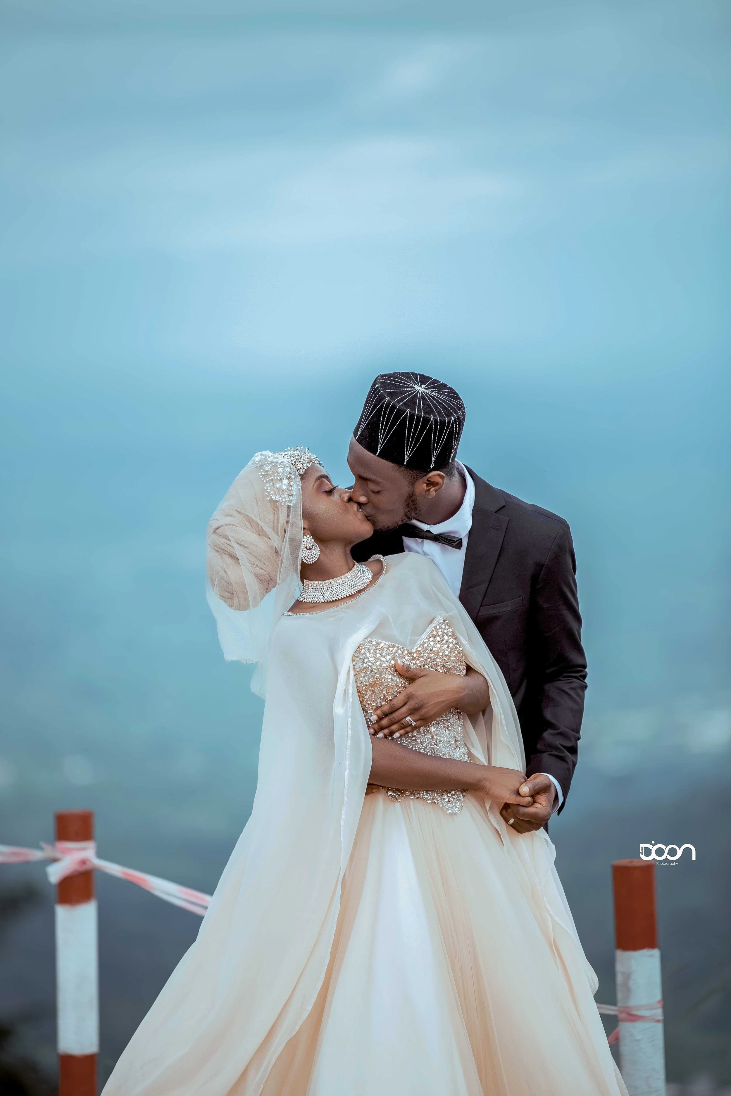 A bride and groom sharing a kiss outdoors, dressed in wedding attire with a cloudy sky in the background.
