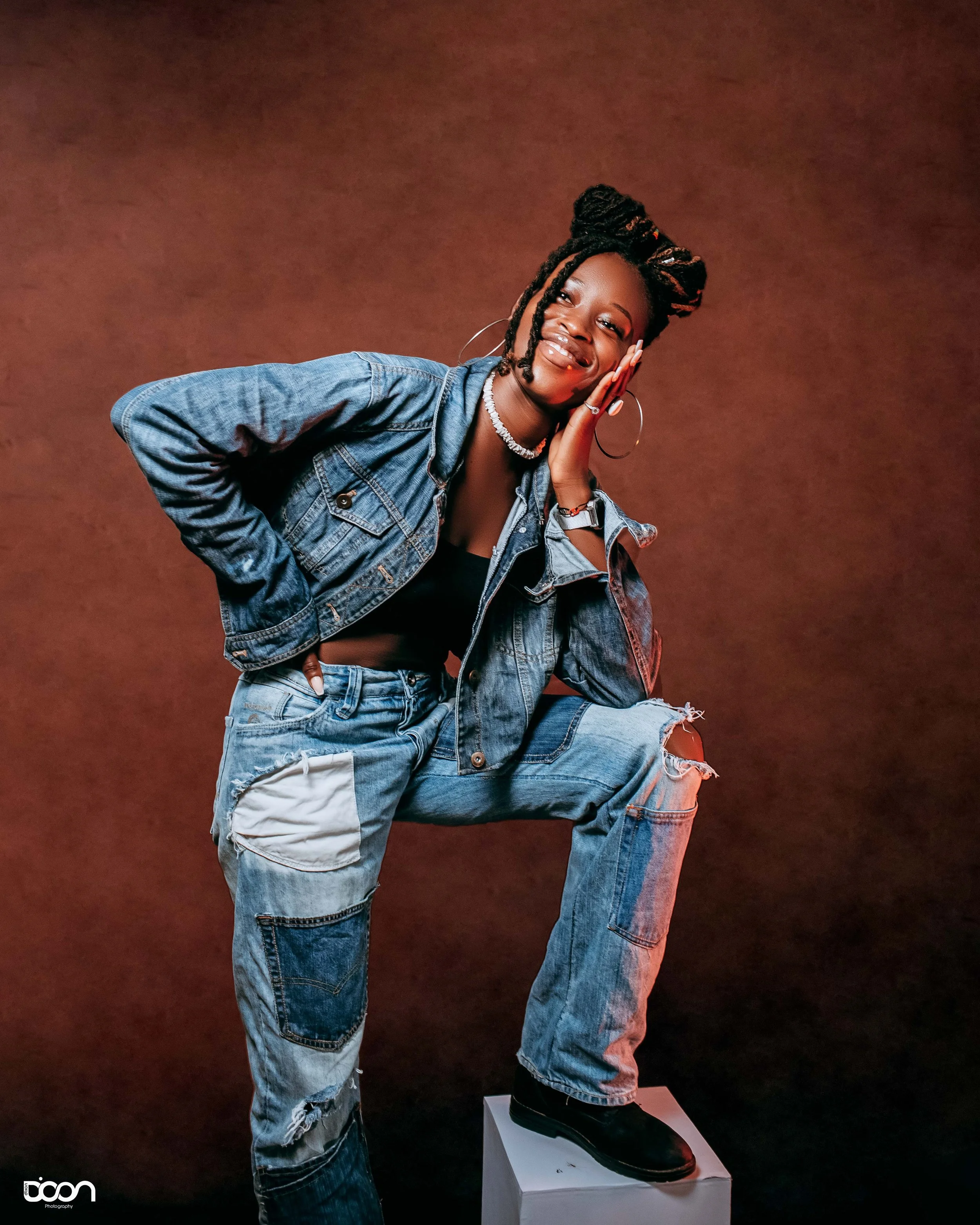 Young woman with dreadlocks in a denim jacket and ripped jeans, smiling, posing against a brown background.