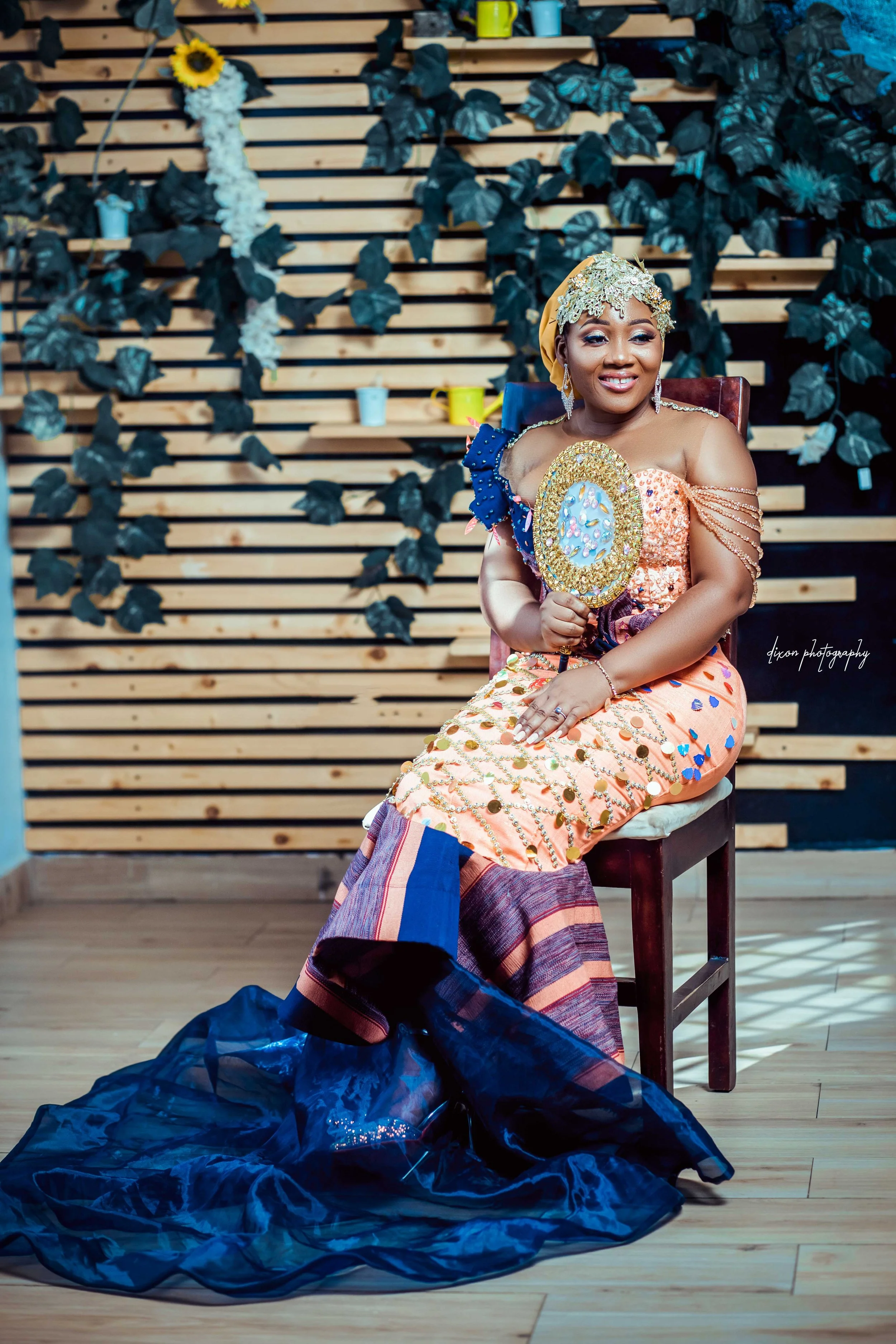A woman dressed in traditional African attire, sitting on a wooden chair, holding a decorated hand fan, with an intricate hairstyle decorated with gold and lace. The background features a wooden slat wall decorated with green leafy plants and small c