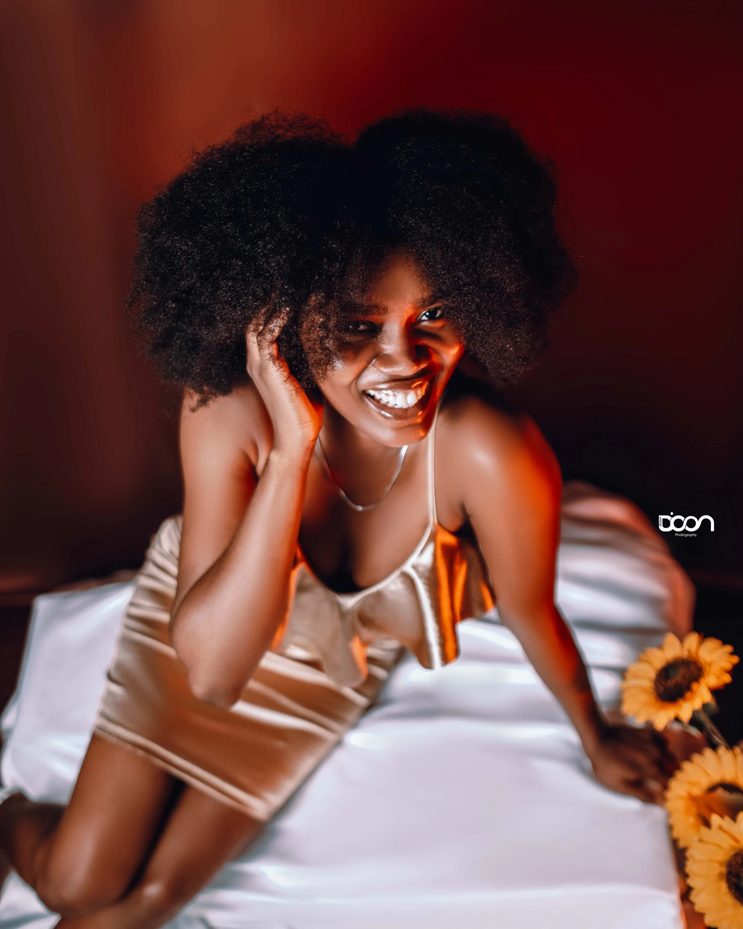 A woman with a big afro hairstyle, smiling and posing on a bed with white sheets, wearing a satin beige-colored dress, with sunflowers beside her and red and orange lighting.