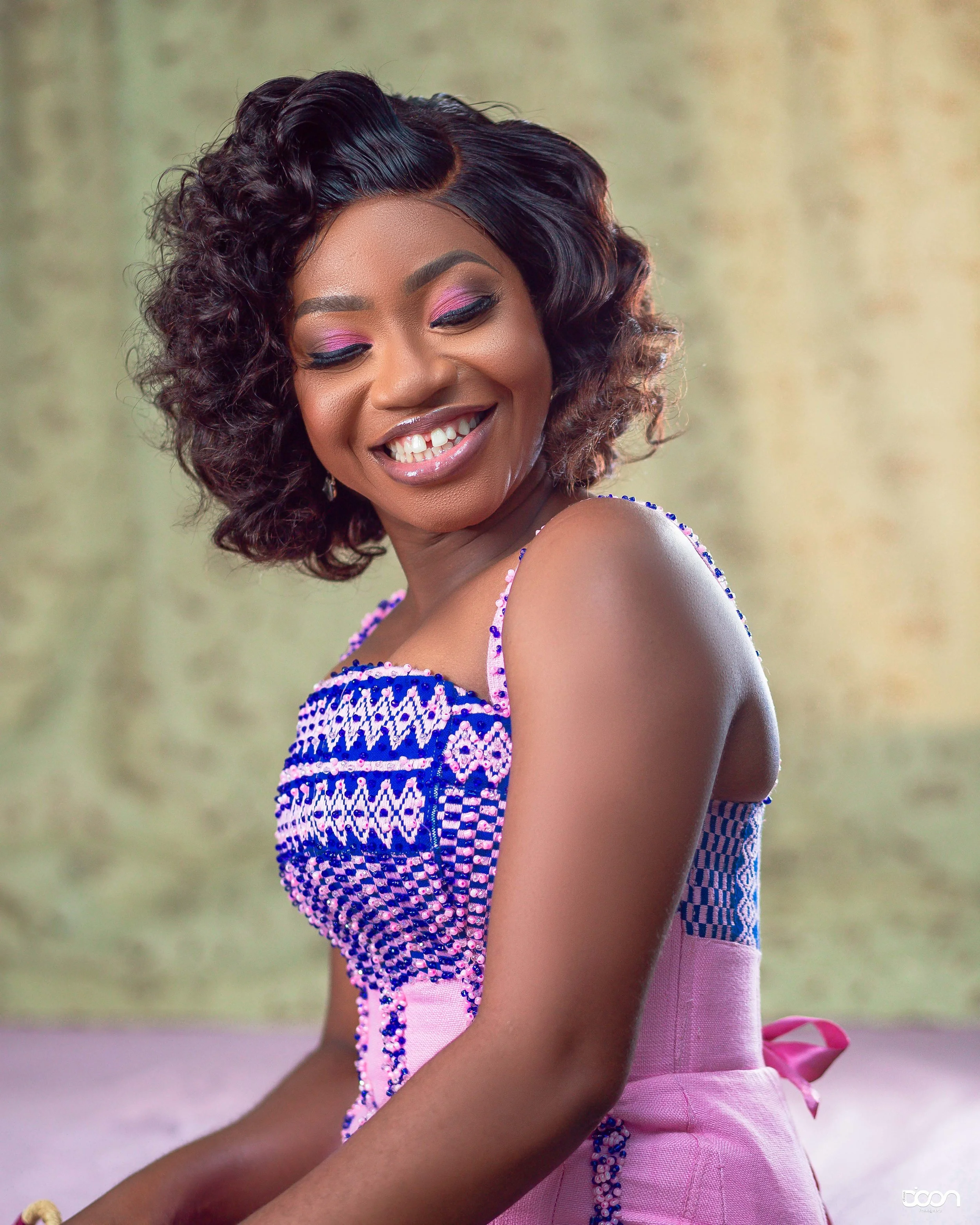 A woman with curly dark hair styled in a vintage bob, smiling and looking down, wearing a colorful beaded dress with pink and purple tones, and a pink sash tied in a bow at her waist, set against a neutral background.