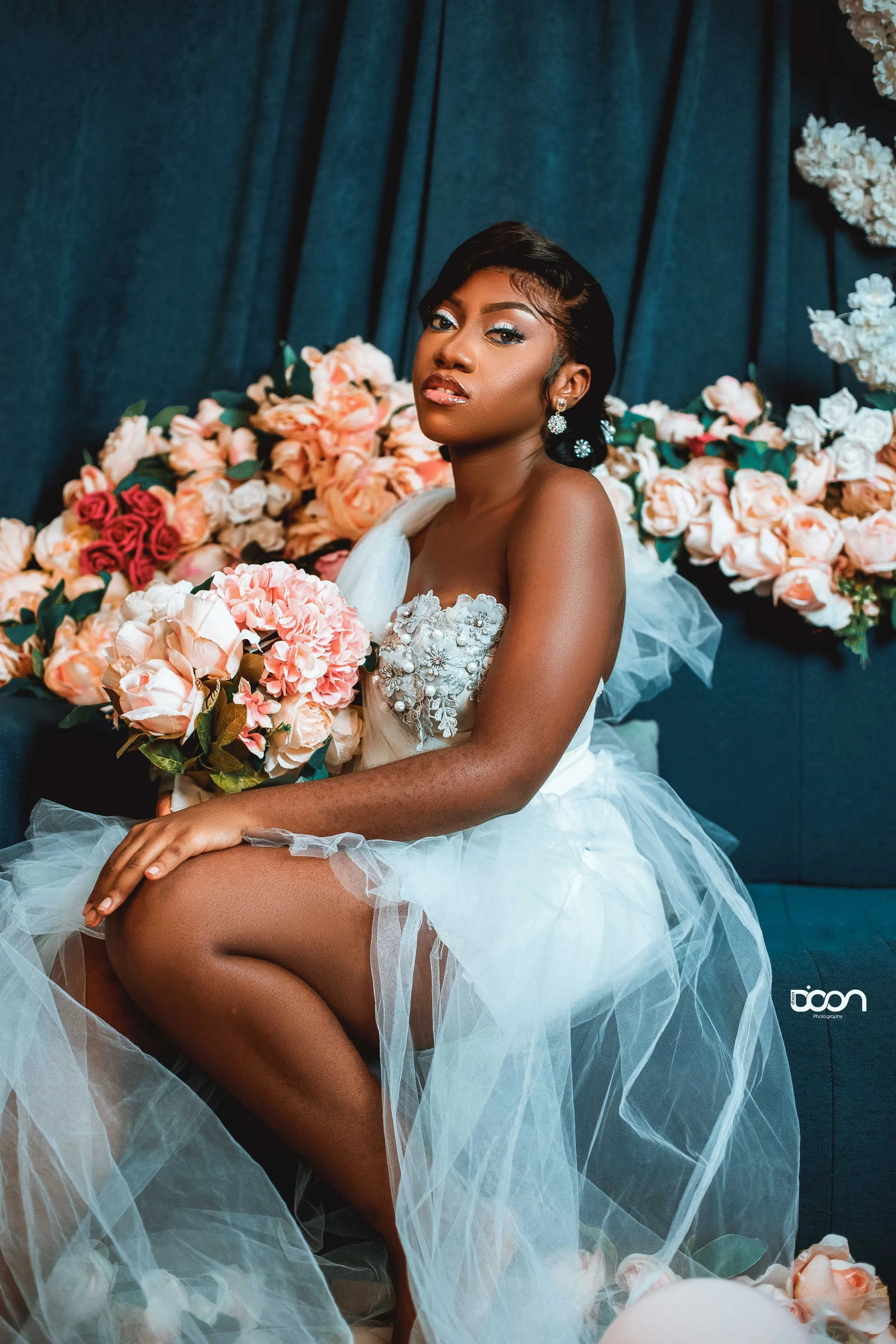 A woman in a wedding dress sitting on a blue couch surrounded by pink and white roses. She has short hair, earrings, and is holding a bouquet of pink and white flowers.