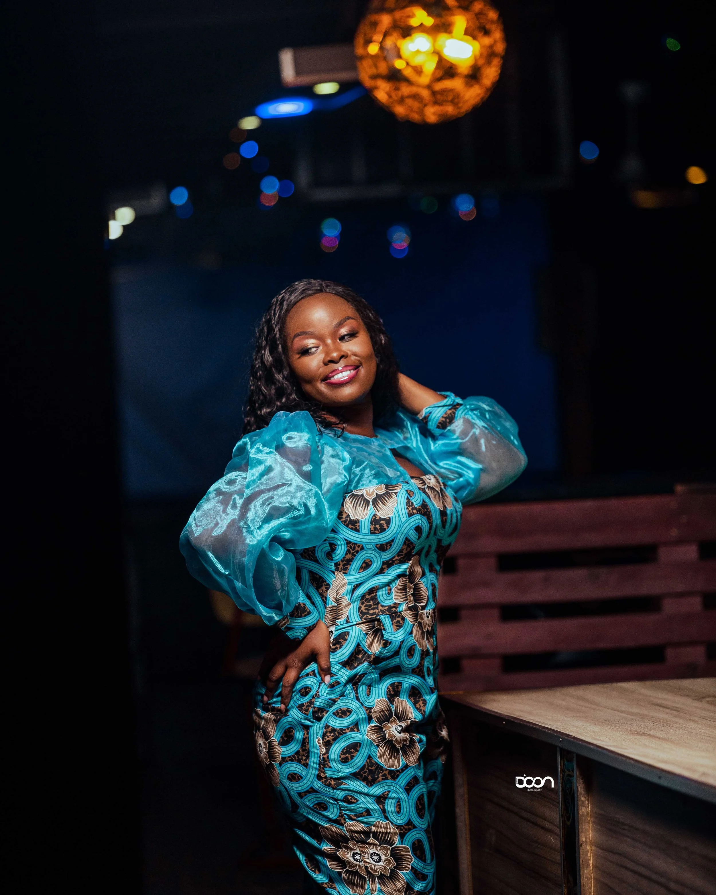 A woman in a vibrant, colorful dress with blue and brown floral patterns, standing indoors near a wooden counter, smiling and posing with one hand on her hip, in a dimly lit setting with bokeh lights in the background.
