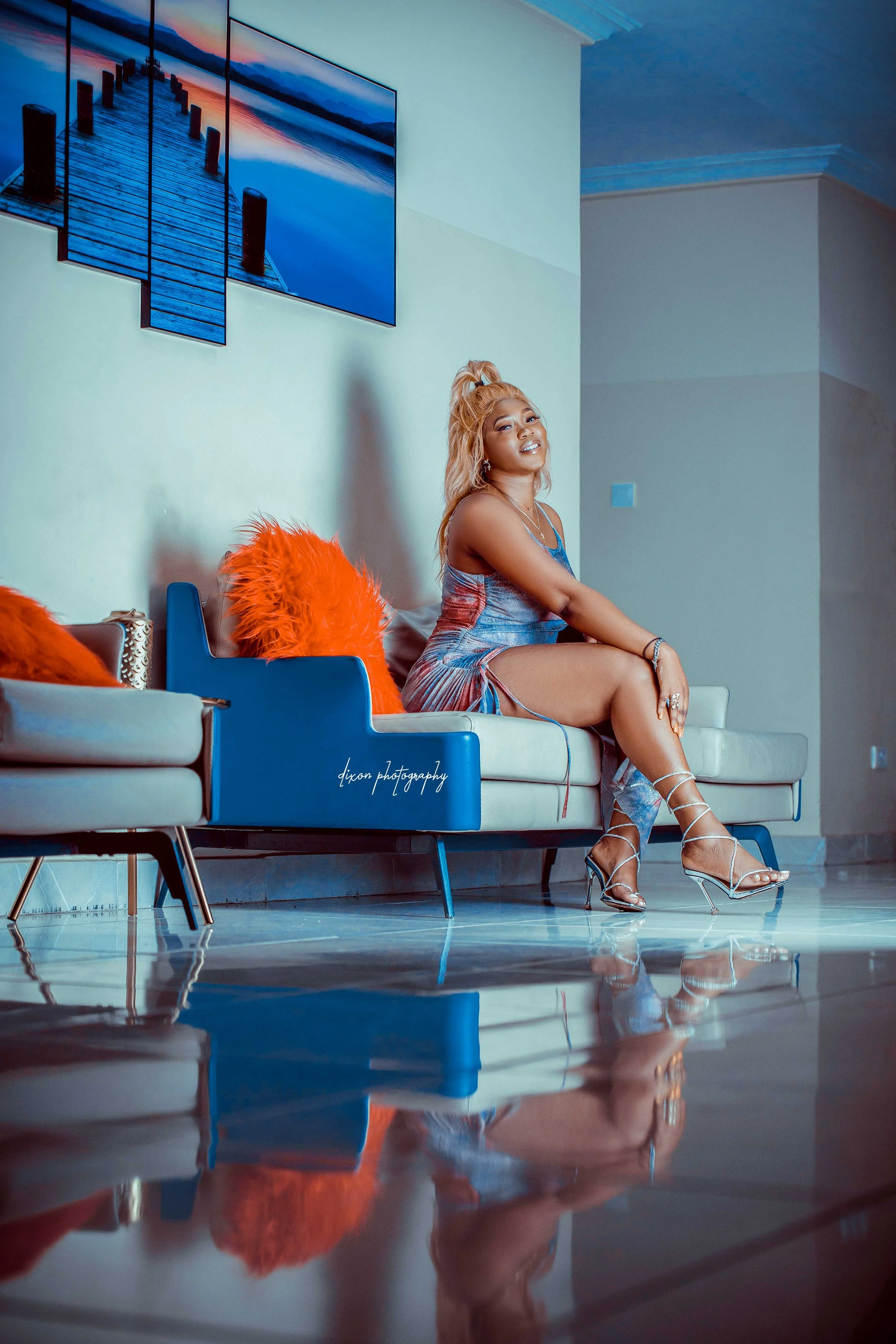 A woman with long wavy blonde hair in a high ponytail, wearing a shiny multicolored dress with side slits, high heels, and jewelry, sits on a modern sofa with orange fuzzy pillows, smiling at the camera. A large multi-paneled wall art of a dock exten