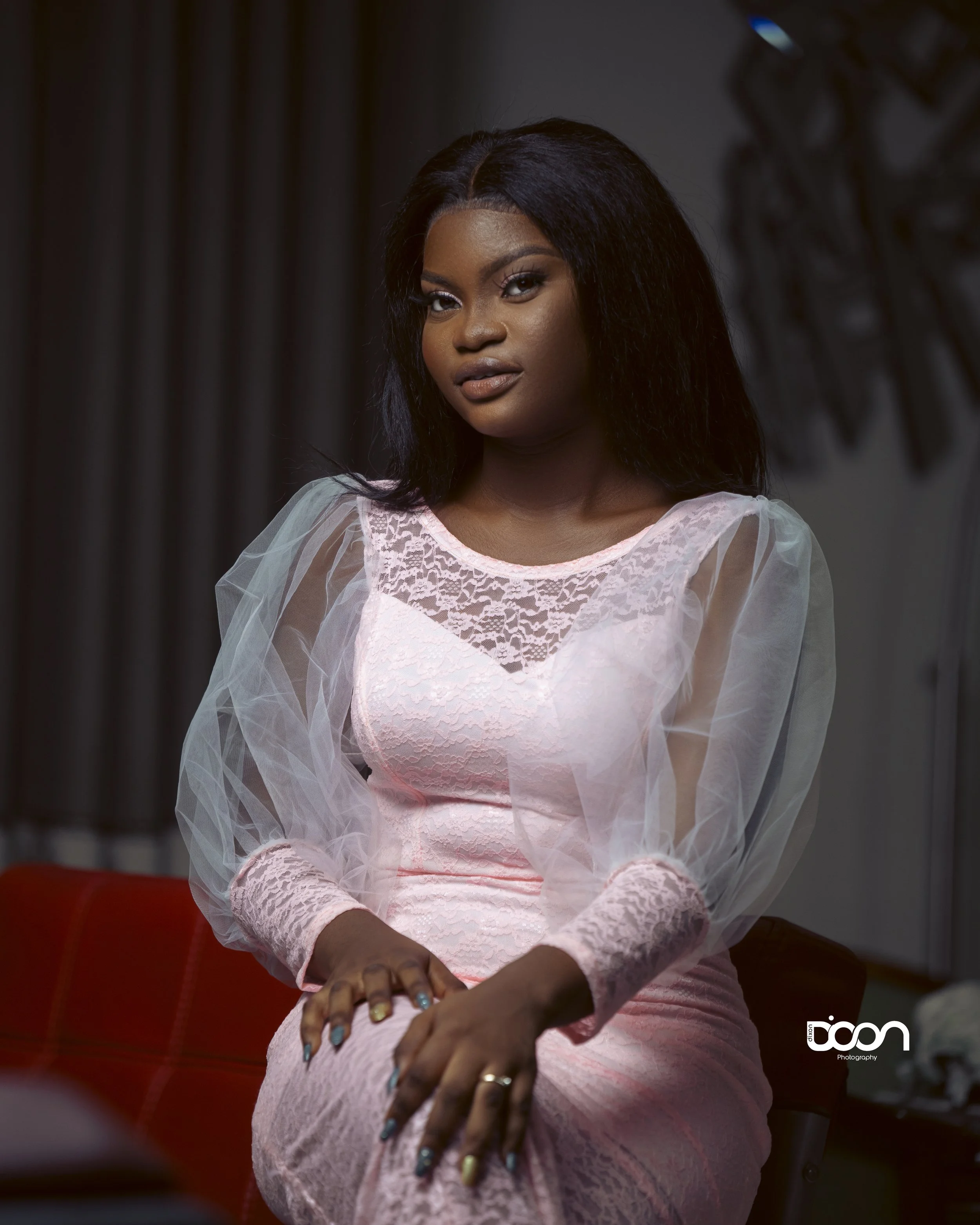A woman with dark hair wearing a pink lace dress and sheer long sleeves, sitting on a red chair in a dimly lit room.
