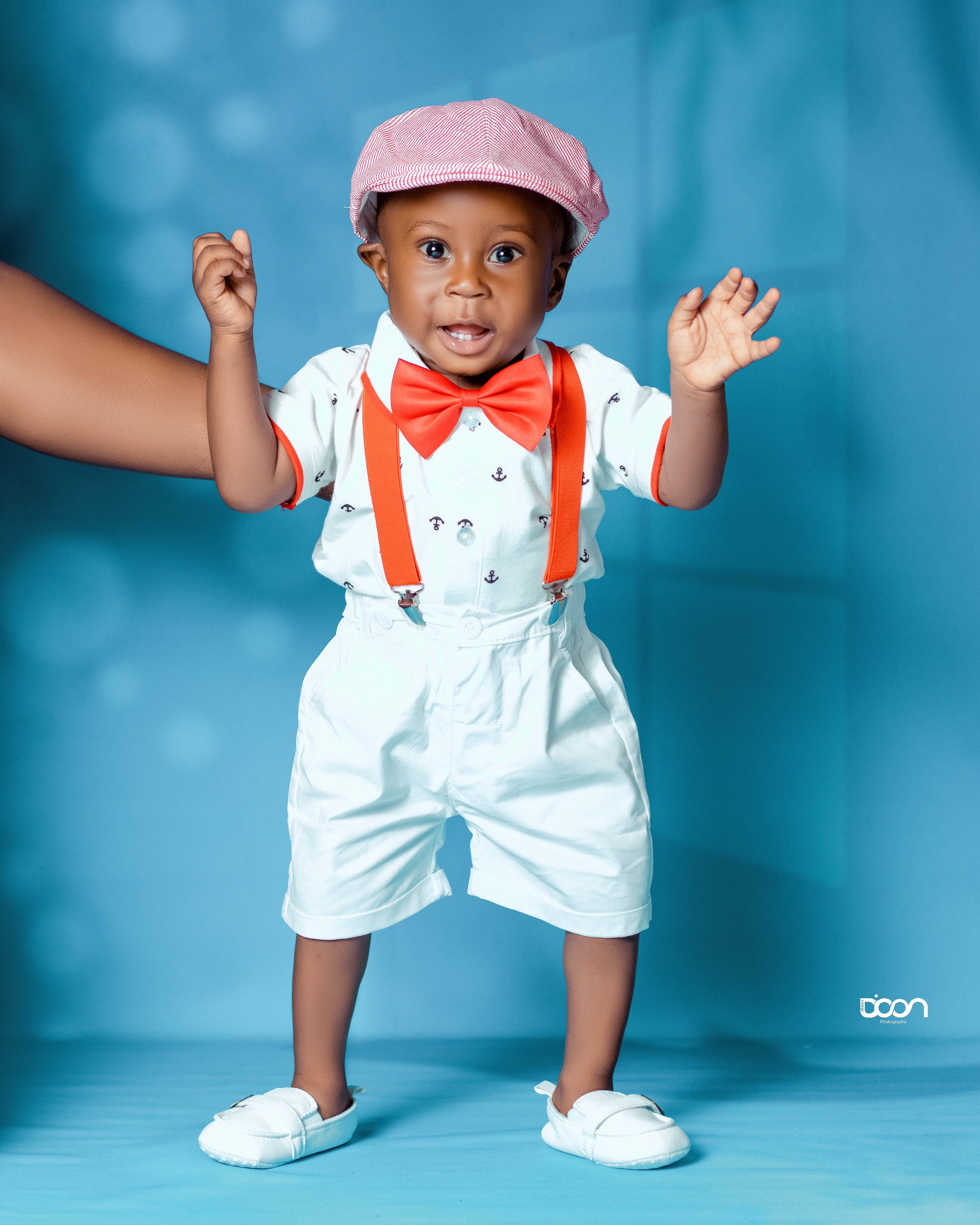 A young boy dressed in white shorts, a white shirt with small anchor patterns, orange suspenders, a red bow tie, and a pink striped cap, standing against a blue background.