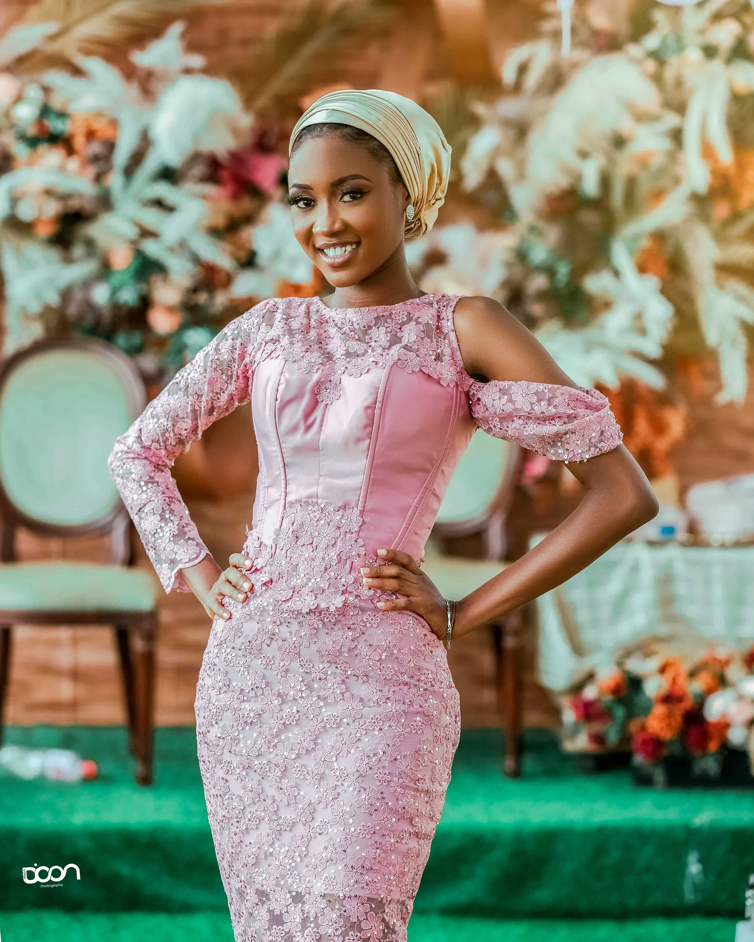 A woman wearing a pink lace dress with off-shoulder sleeves and a matching headwrap, standing confidently with her hands on her hips, in front of a floral backdrop at an indoor event.