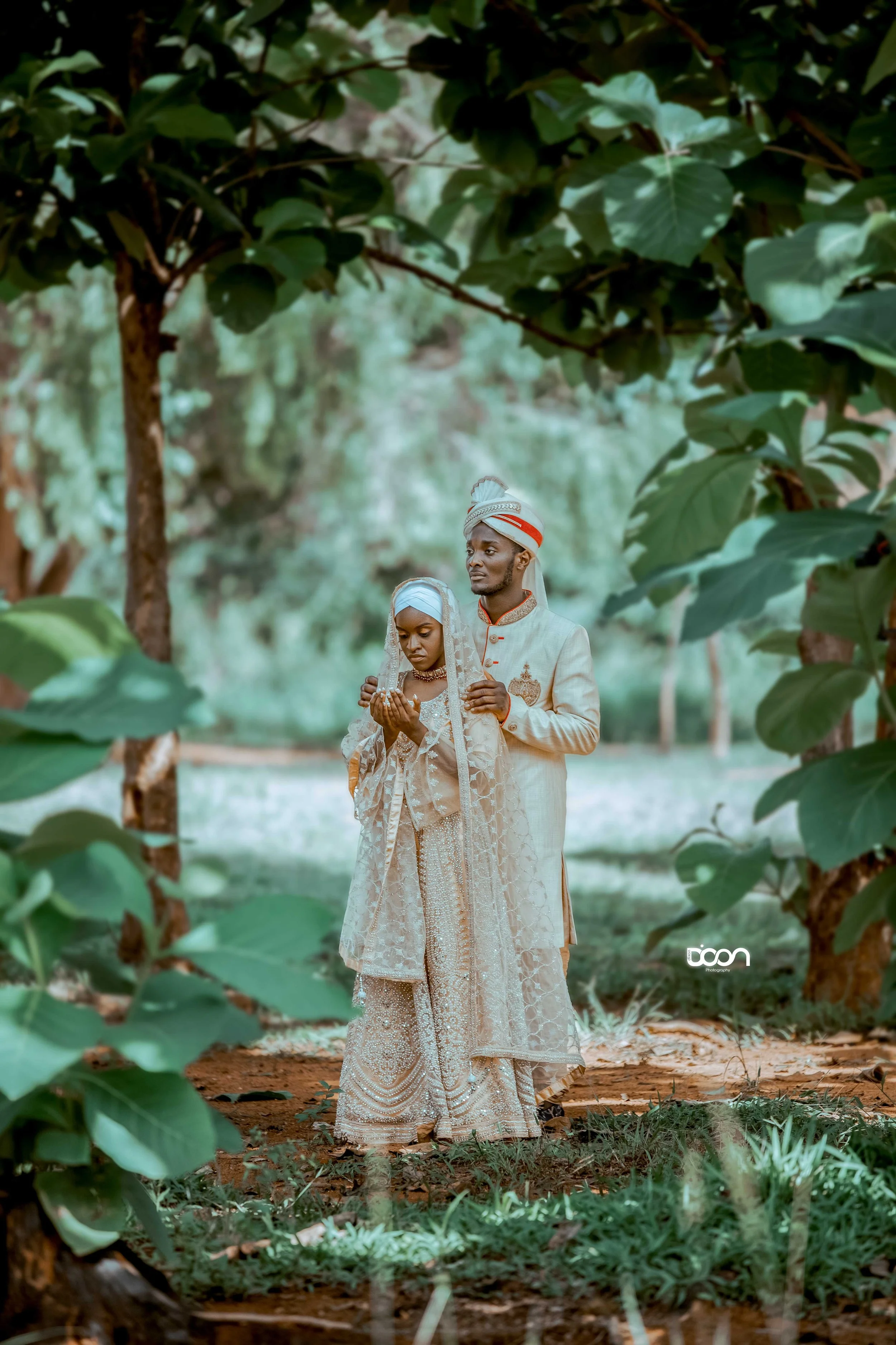 A bride and groom dressed in traditional attire standing outdoors amidst trees, with the bride holding her hands in prayer and the groom gently holding her shoulders.