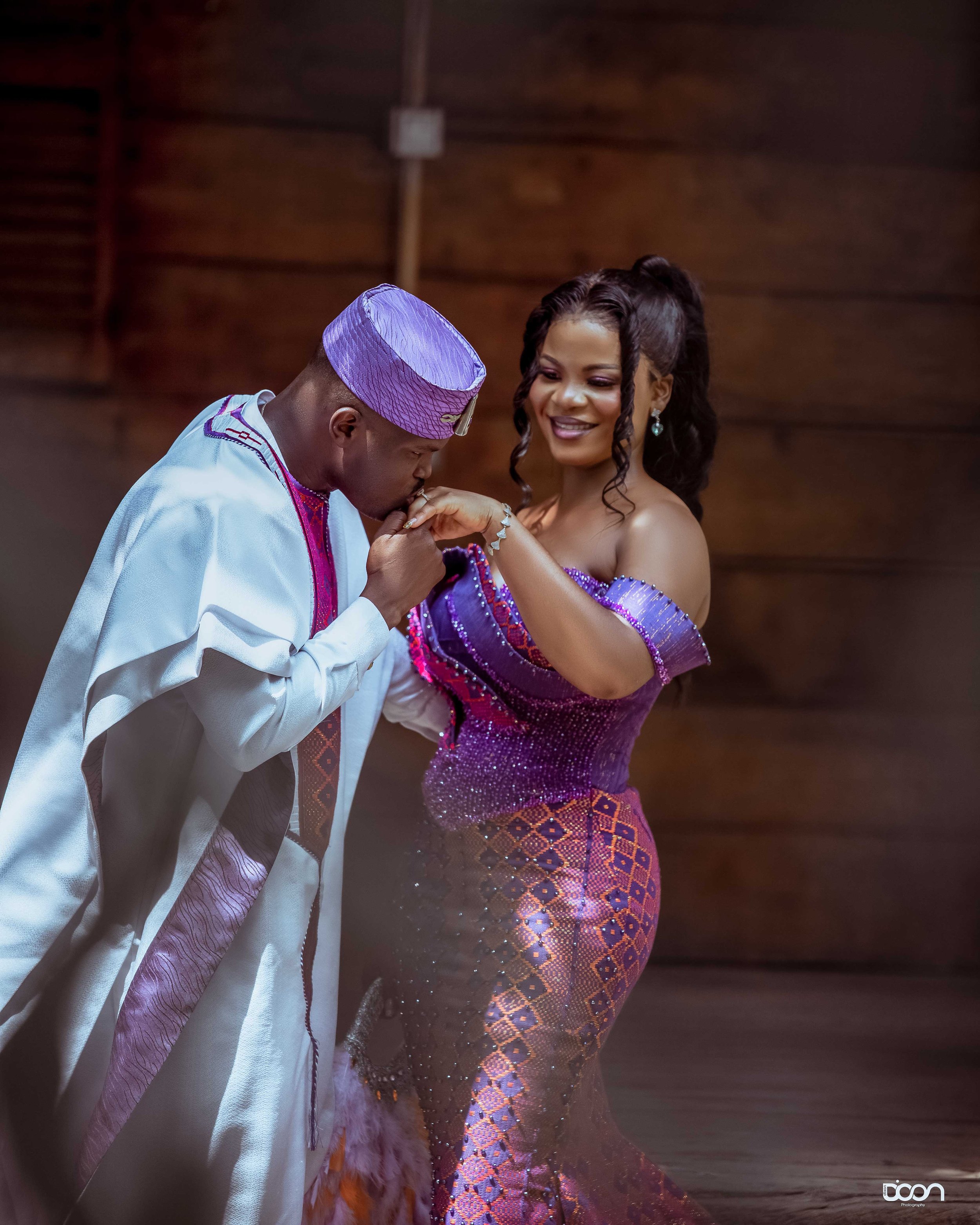 A man kissing a woman's hand during a traditional ceremony, both dressed in vibrant, elaborate African attire, with a wooden background.