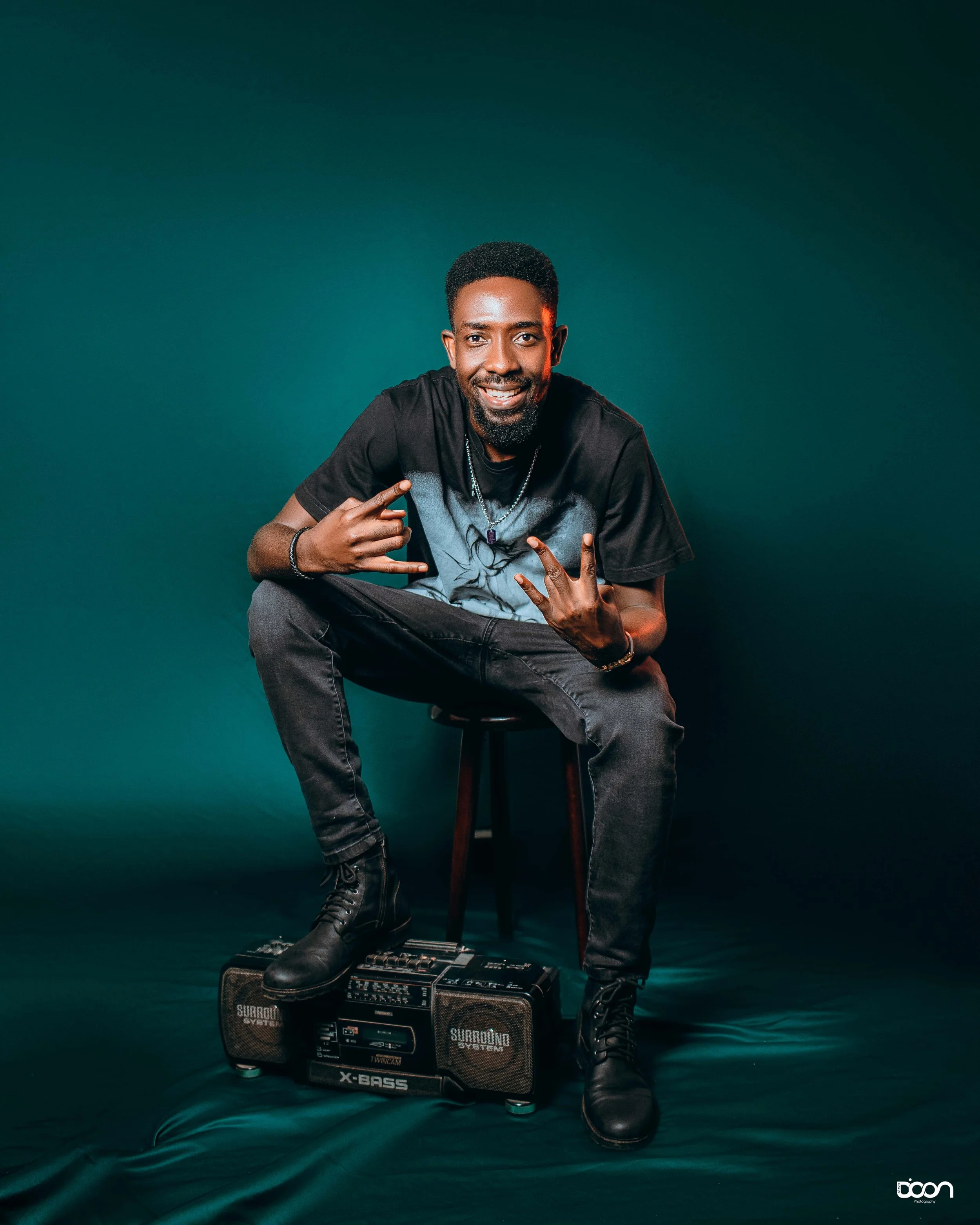 A man sitting on a chair with one leg propped up on a portable stereo, smiling and making hand gestures, in front of a dark green background.