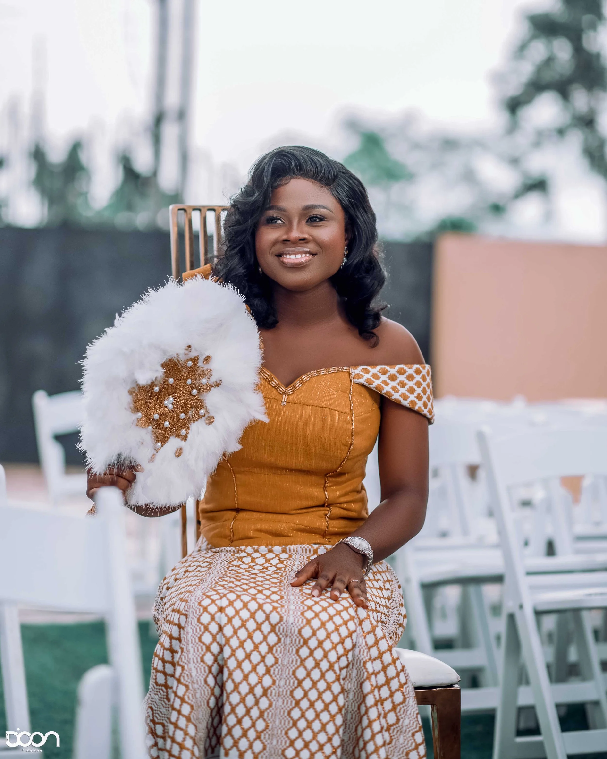 A woman in an orange dress sitting on a chair holding a white feathered fan with brown embellishments, outdoors with white chairs in the background.