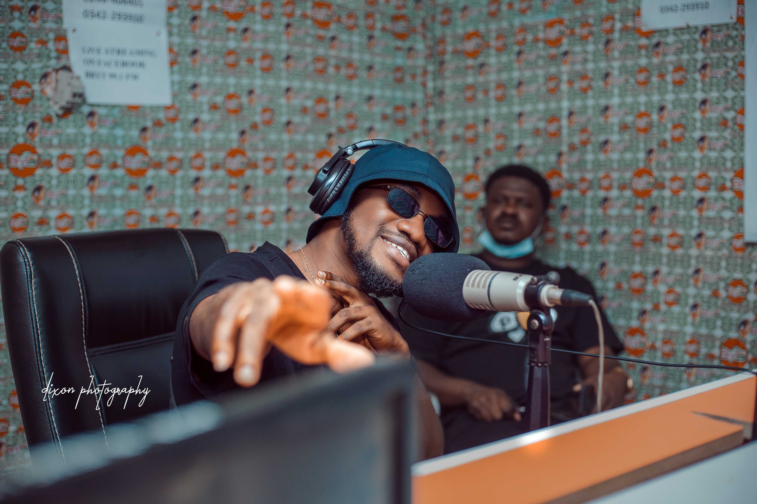 A man wearing sunglasses, a blue cap, and headphones smiling and pointing at the camera while speaking into a microphone in a radio station studio, with another man in the background wearing a mask around his neck.