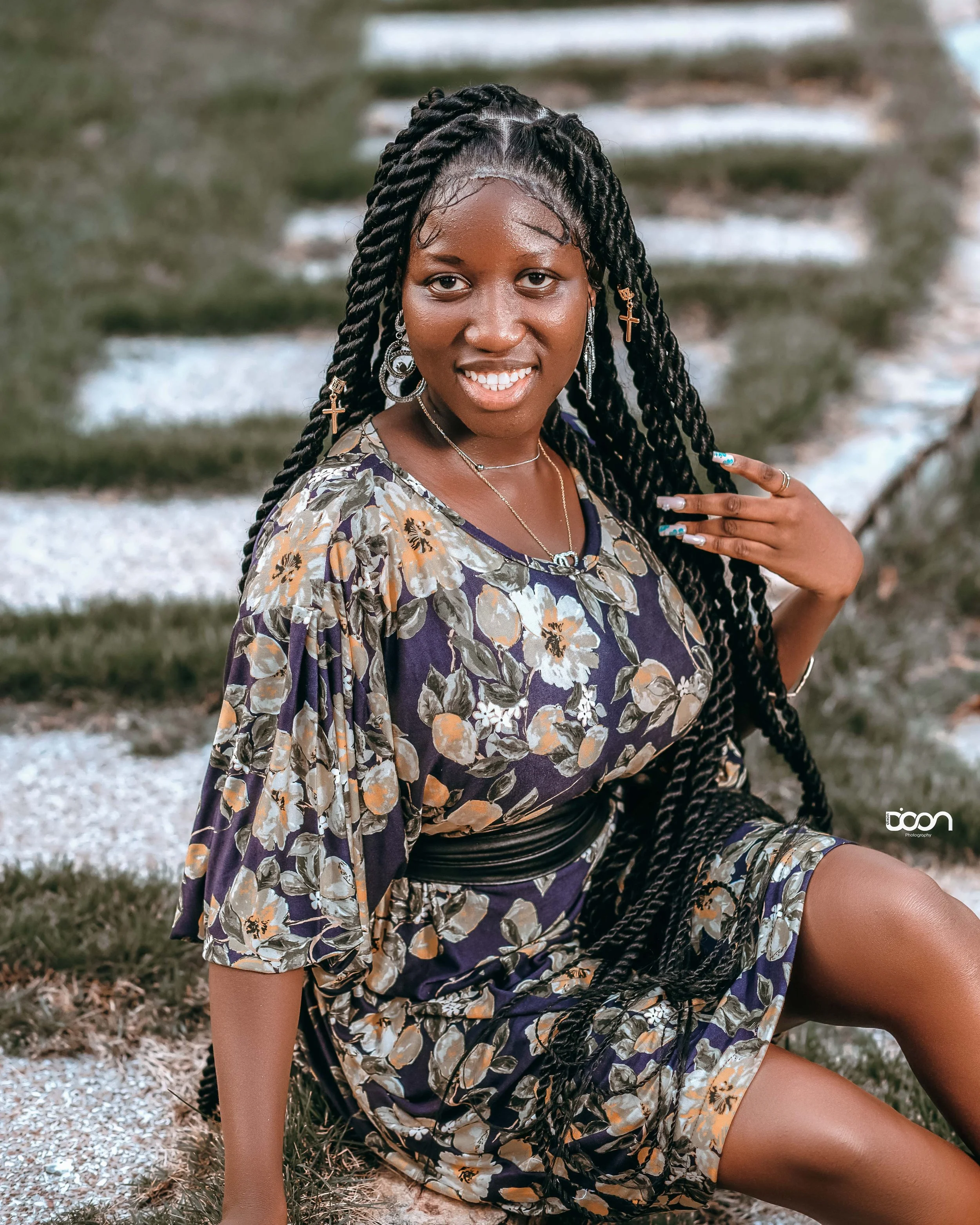 A woman sitting on grass with stone pathway behind her, wearing a floral dress and jewelry, smiling at the camera.