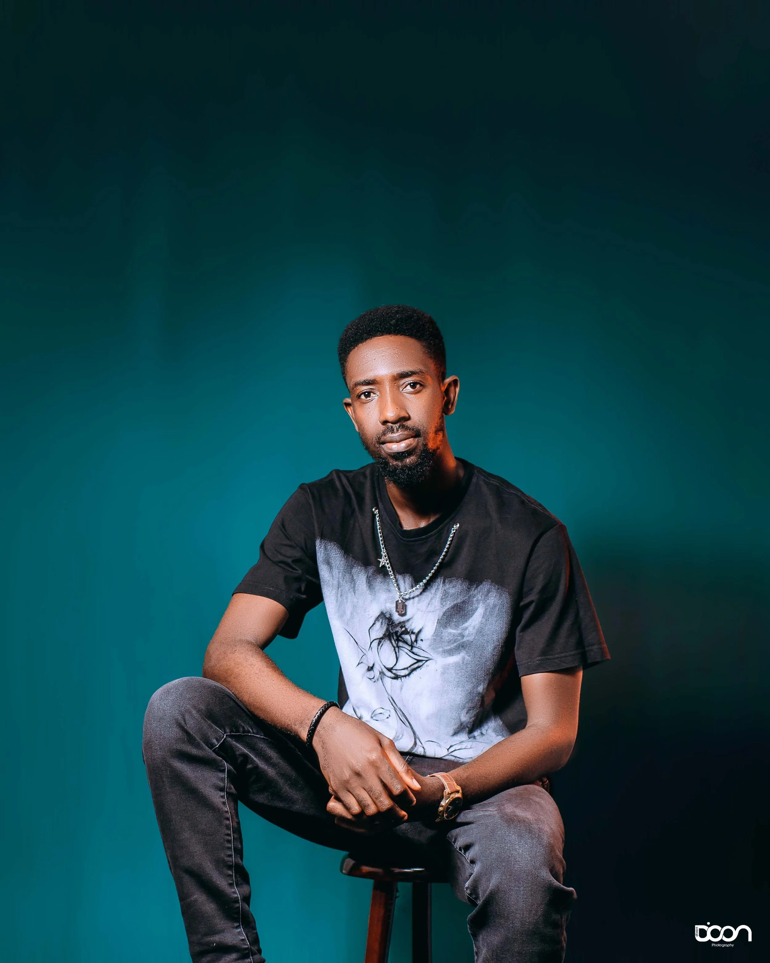 A young man with dark skin, short curly hair, and a beard, sitting on a stool against a dark teal background. He is wearing a black T-shirt with a graphic print, a silver chain necklace, a black bracelet, a gold watch, and dark jeans. He has a seriou