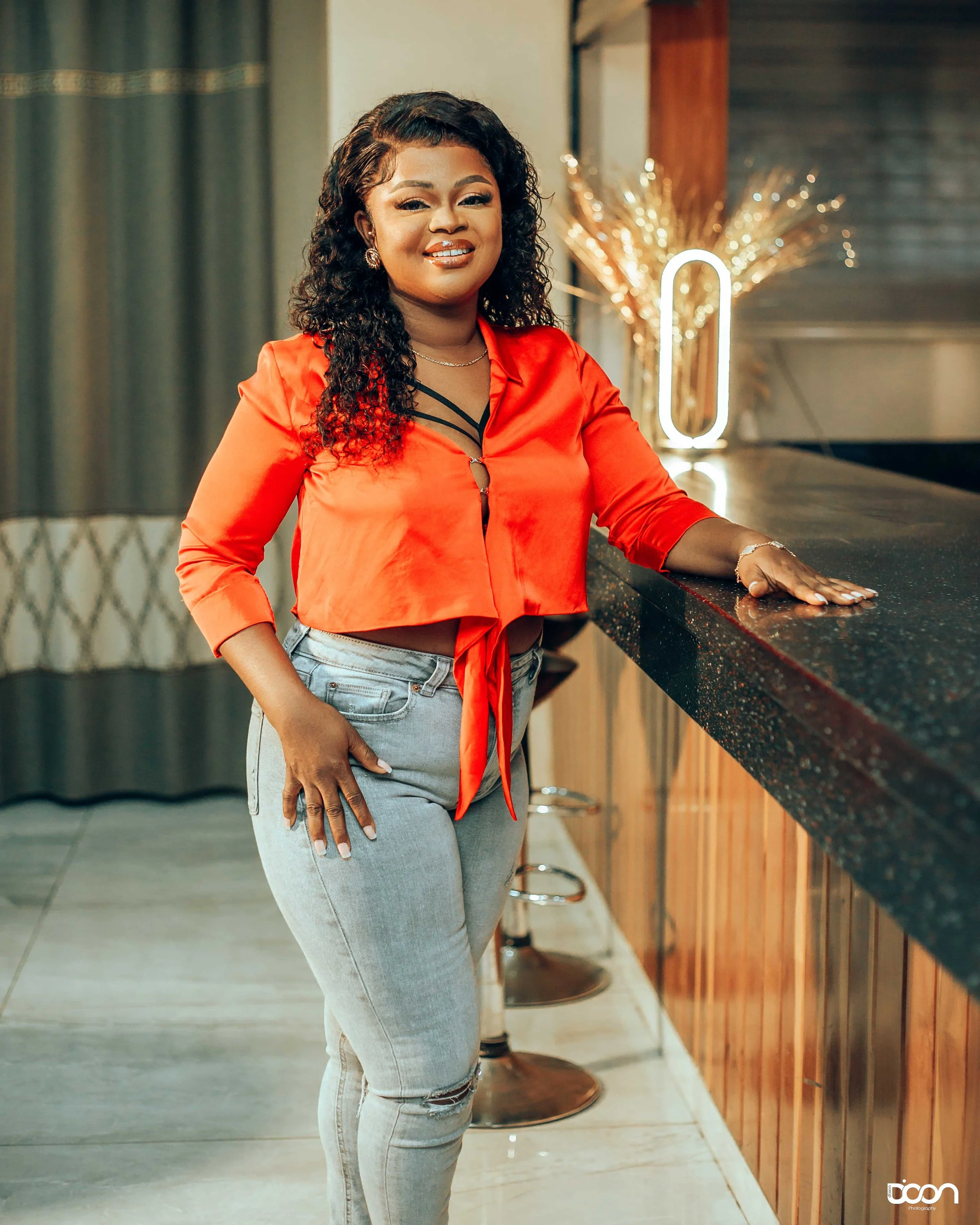 Woman with dark curly hair wearing a red cropped top with black straps and light wash jeans, standing at a bar counter in a modern room, smiling at the camera.