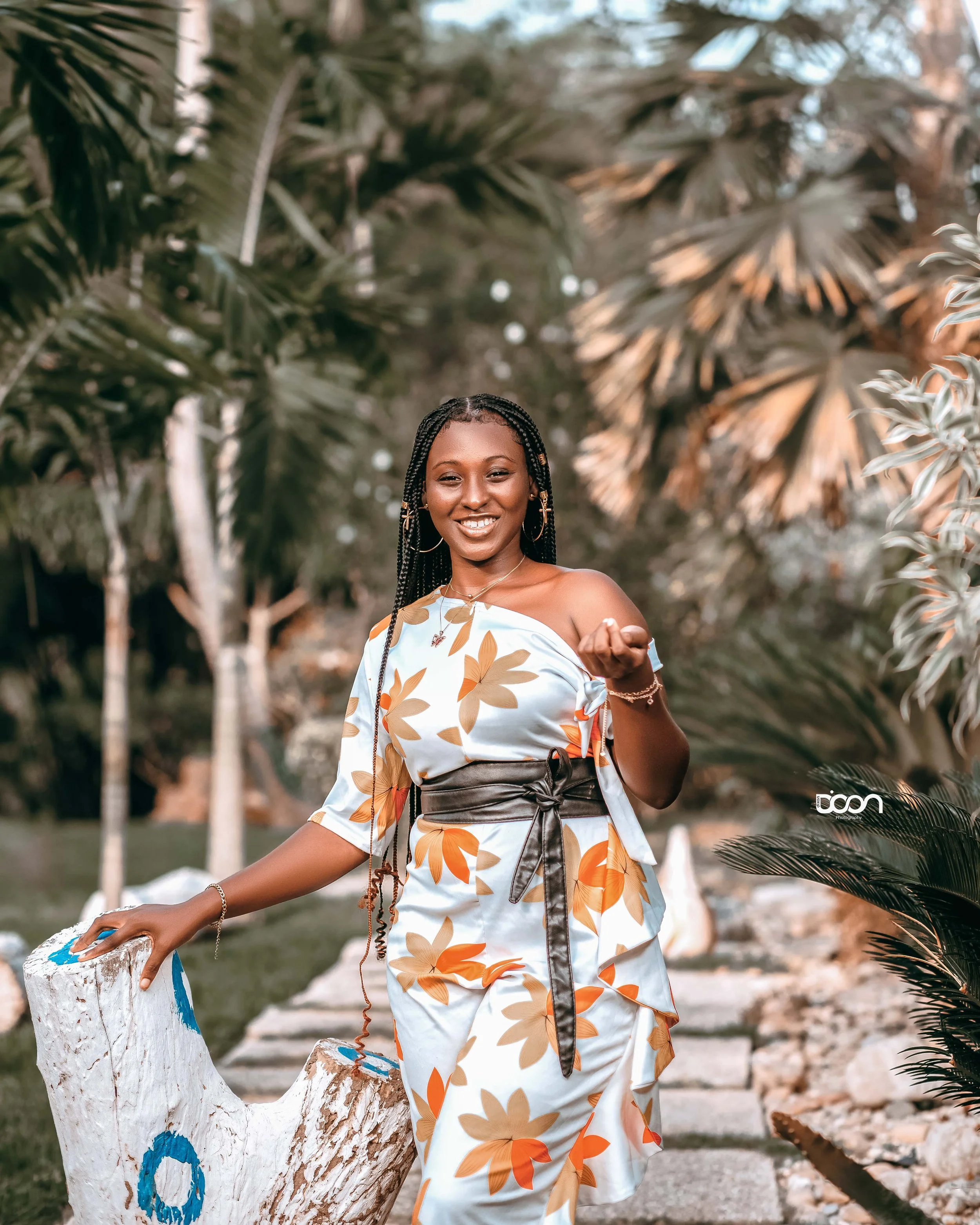 A woman in a floral dress with a black belt, smiling outdoors surrounded by palm trees.