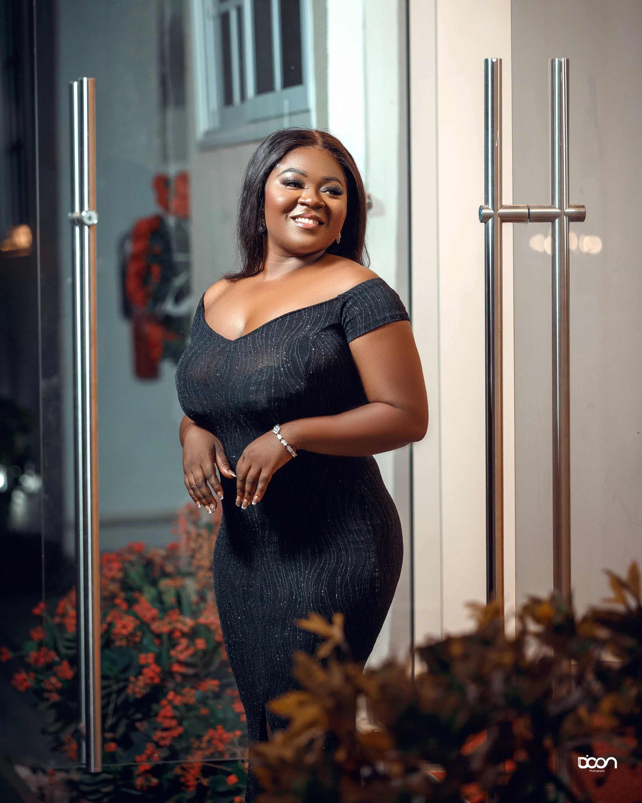 A woman with dark hair wearing a fitted black dress standing near a glass door, smiling with plants in the foreground.