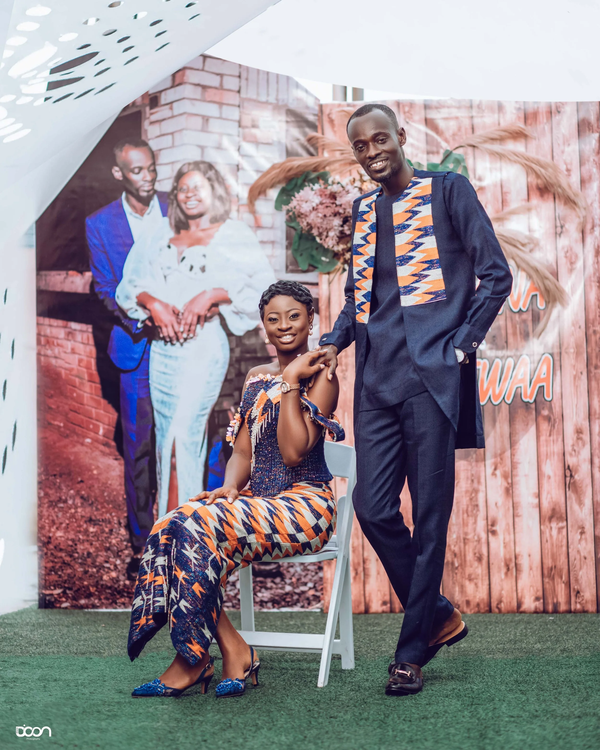 A man and woman in traditional African attire posing at a photo shoot with a backdrop. The woman is seated on a chair, smiling, and the man is standing beside her, holding her hand and smiling.