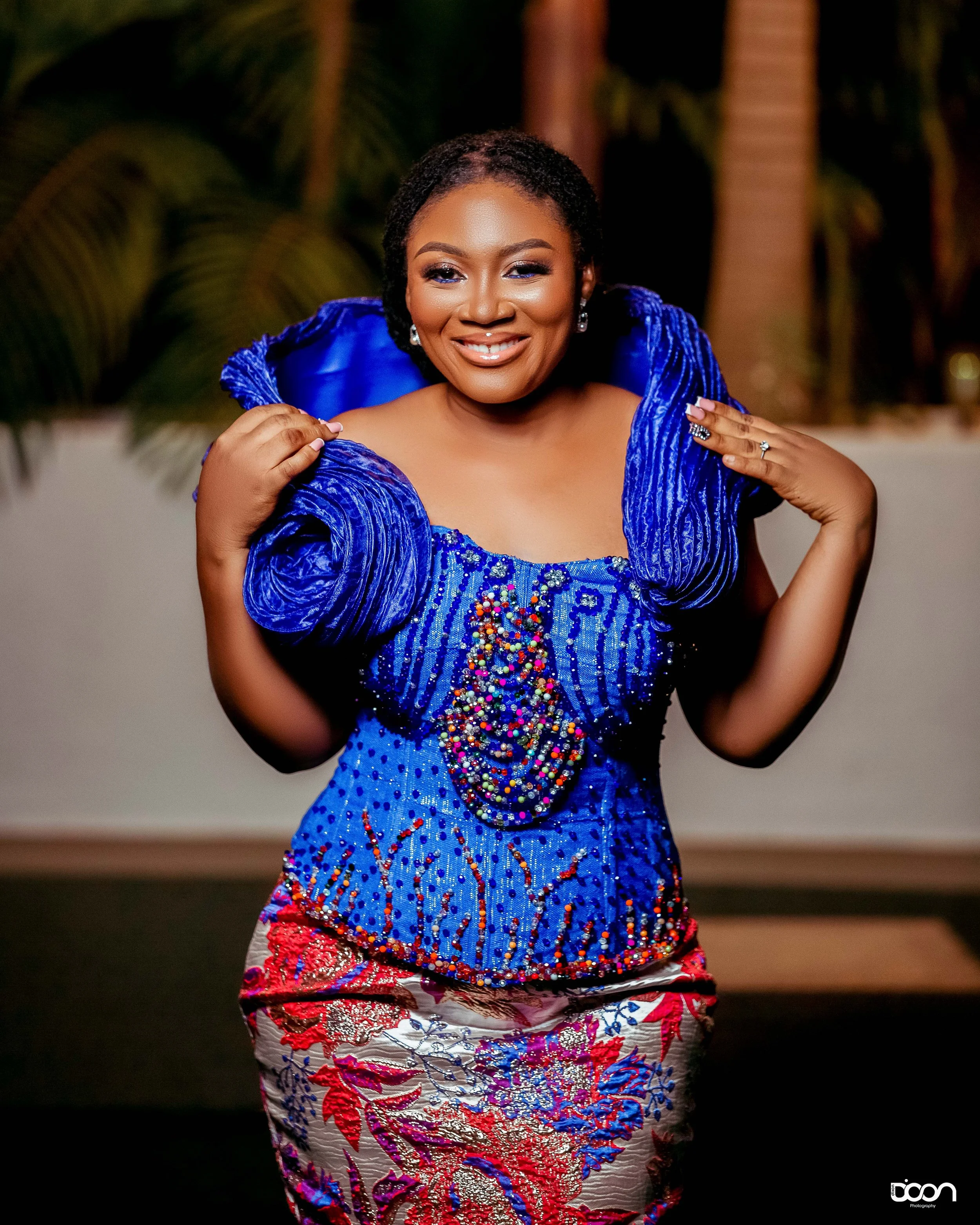 A woman in a vibrant, blue traditional dress adorned with colorful beads, smiling, with palm trees in the background.
