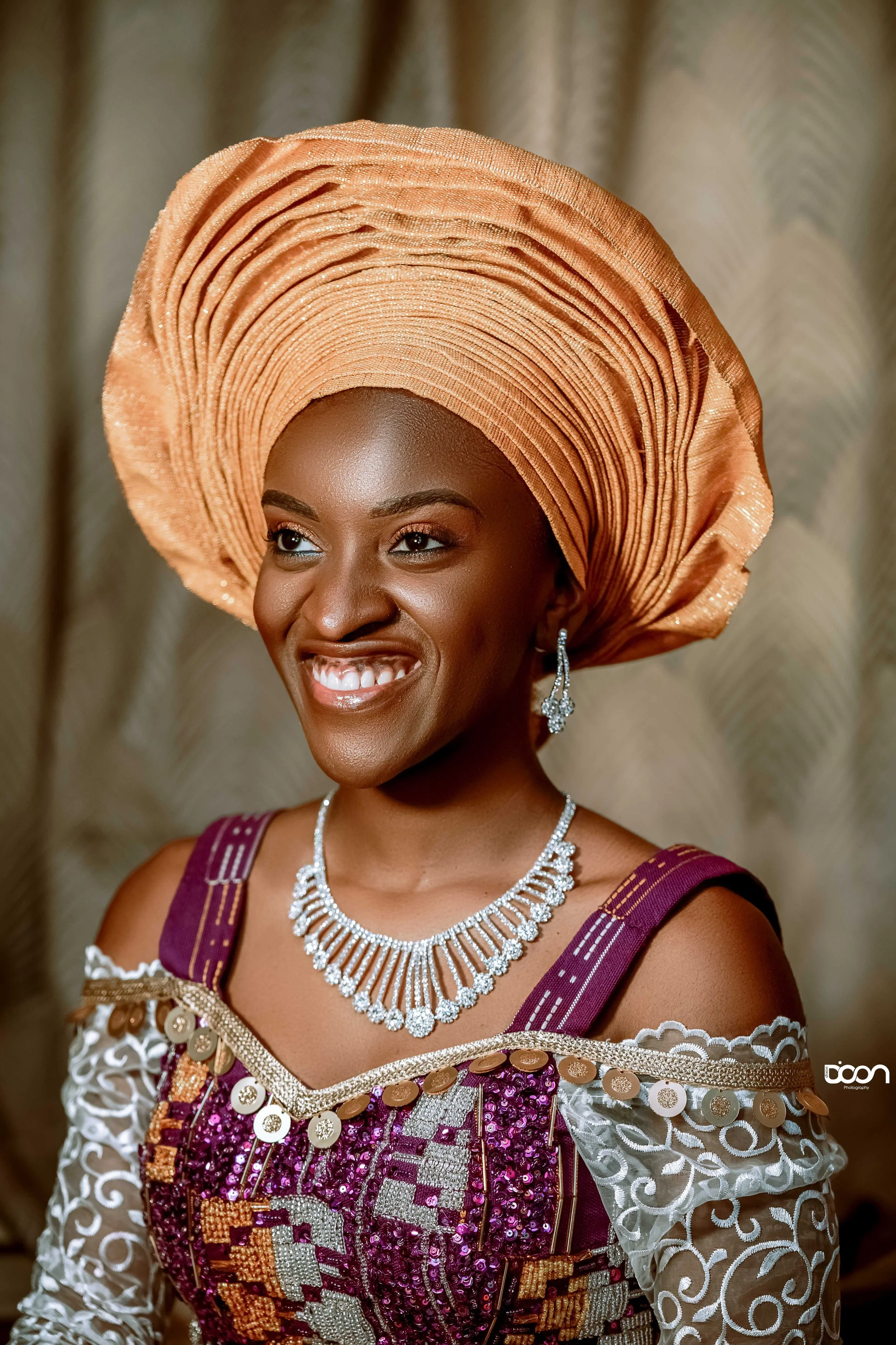 A woman dressed in traditional Nigerian attire, wearing a large peach-colored gele headwrap, ornate jewelry, and a richly decorated purple and gold dress, smiling happily.