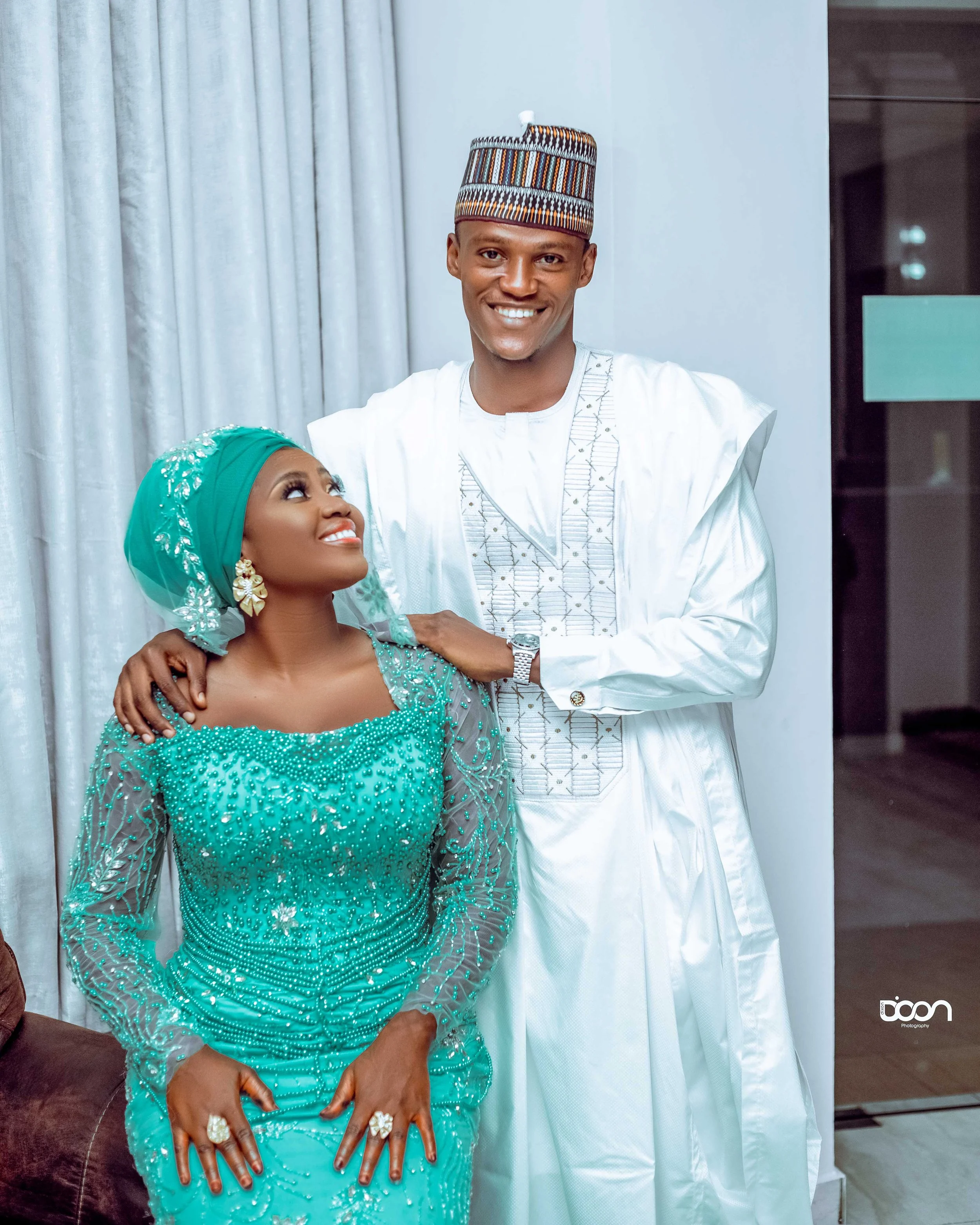 A smiling couple, dressed in traditional Nigerian attire, stand close together indoors. The woman wears a vibrant turquoise lace dress with intricate embellishments and a matching headwrap, while the man wears a white agbada with embroidered details 