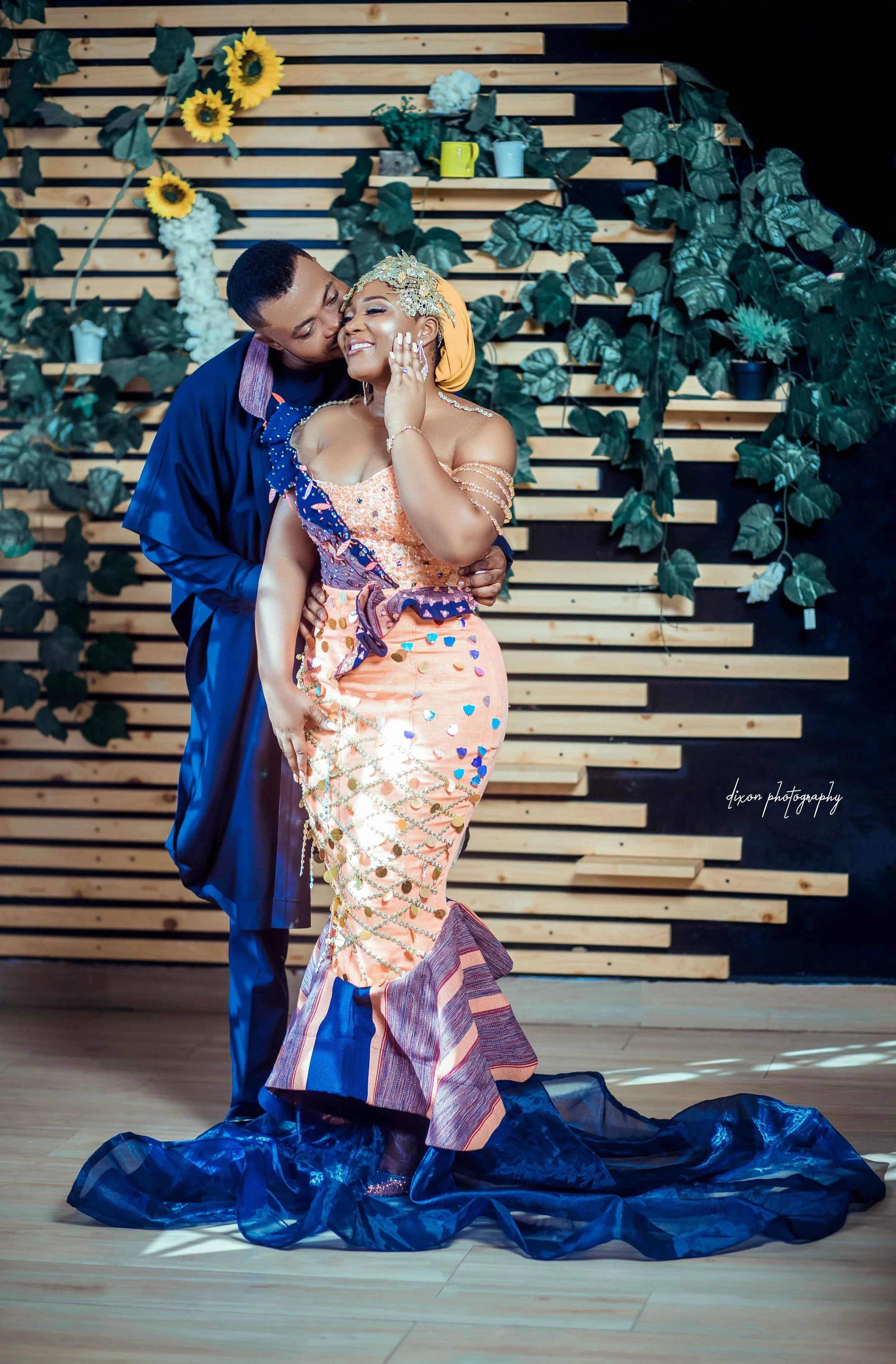 A couple in elegant traditional attire, standing close and smiling affectionately against a decorative backdrop with green leaves and sunflowers. The woman wears a colorful, embellished dress with a train, and the man wears a dark blue outfit, embrac