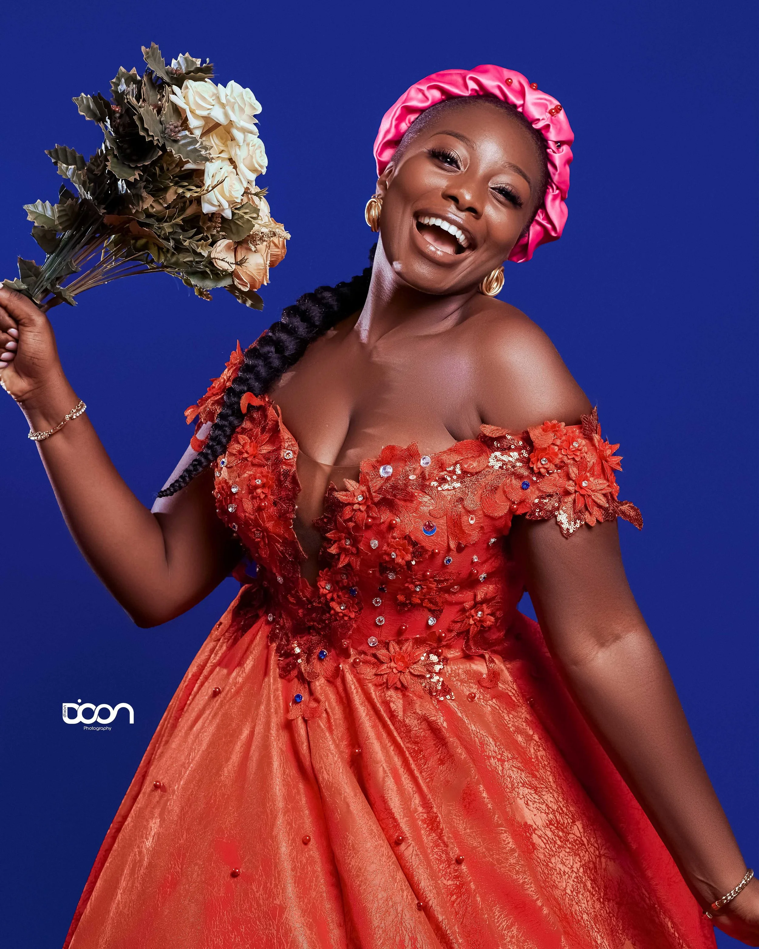 Woman with dark skin smiling, wearing a vibrant red dress with floral details and sequins, pink satin headscarf, holding a bouquet of dried flowers, against a blue background.