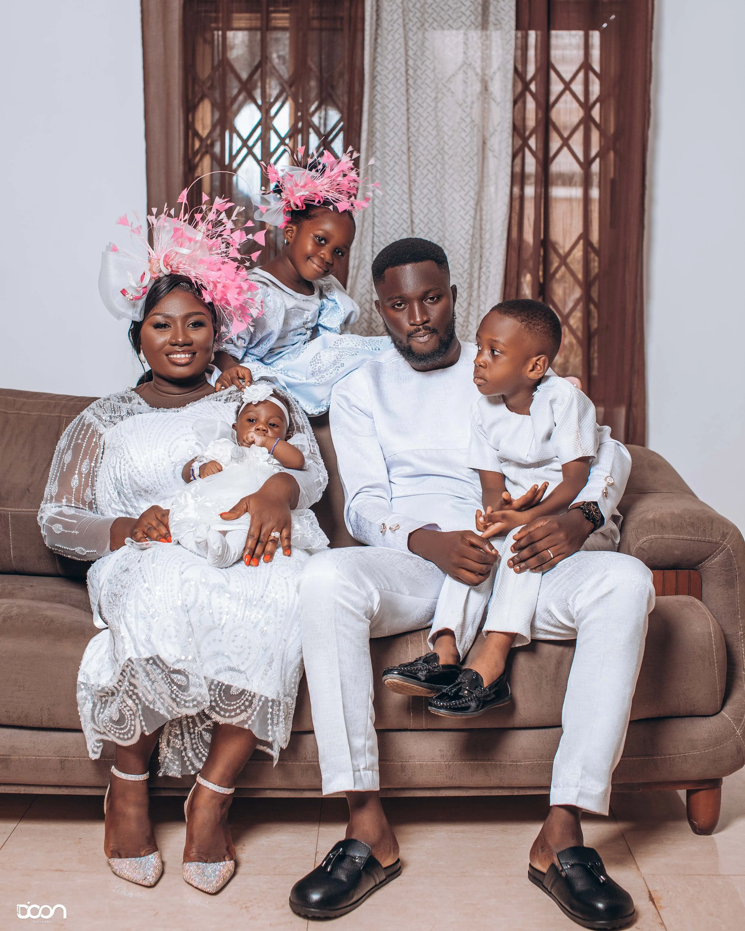 Family of five dressed in white, sitting on a brown sofa. Two young girls wear elaborate pink hats, one young girl is in a white dress and is being held by a woman, and a young boy is sitting on a man's lap. They are indoors with brown lace curtains 