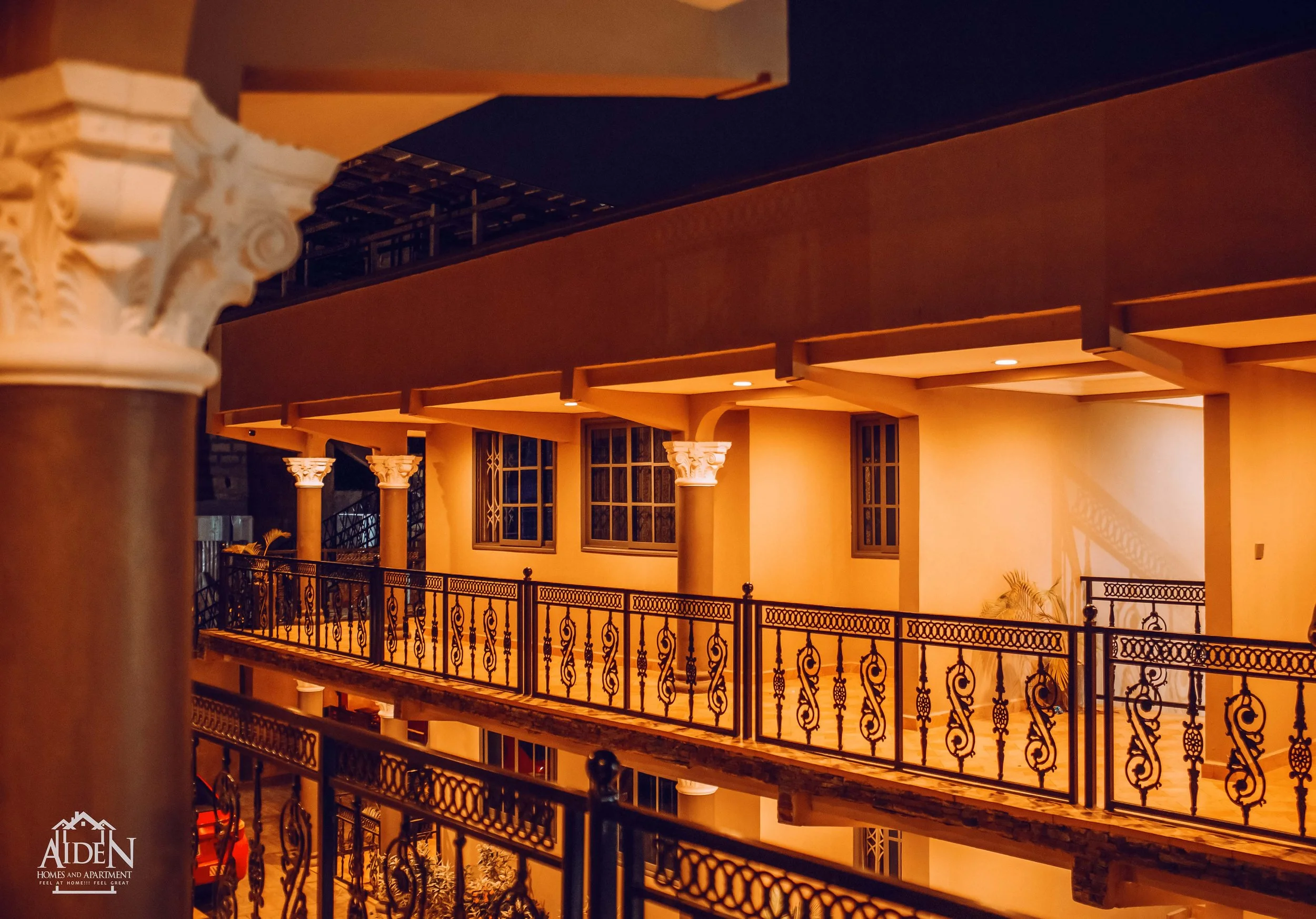 Interior view of a multi-story building with decorative balcony railings, columns with ornate capitals, and warm ambient lighting, possibly a hotel or apartment.